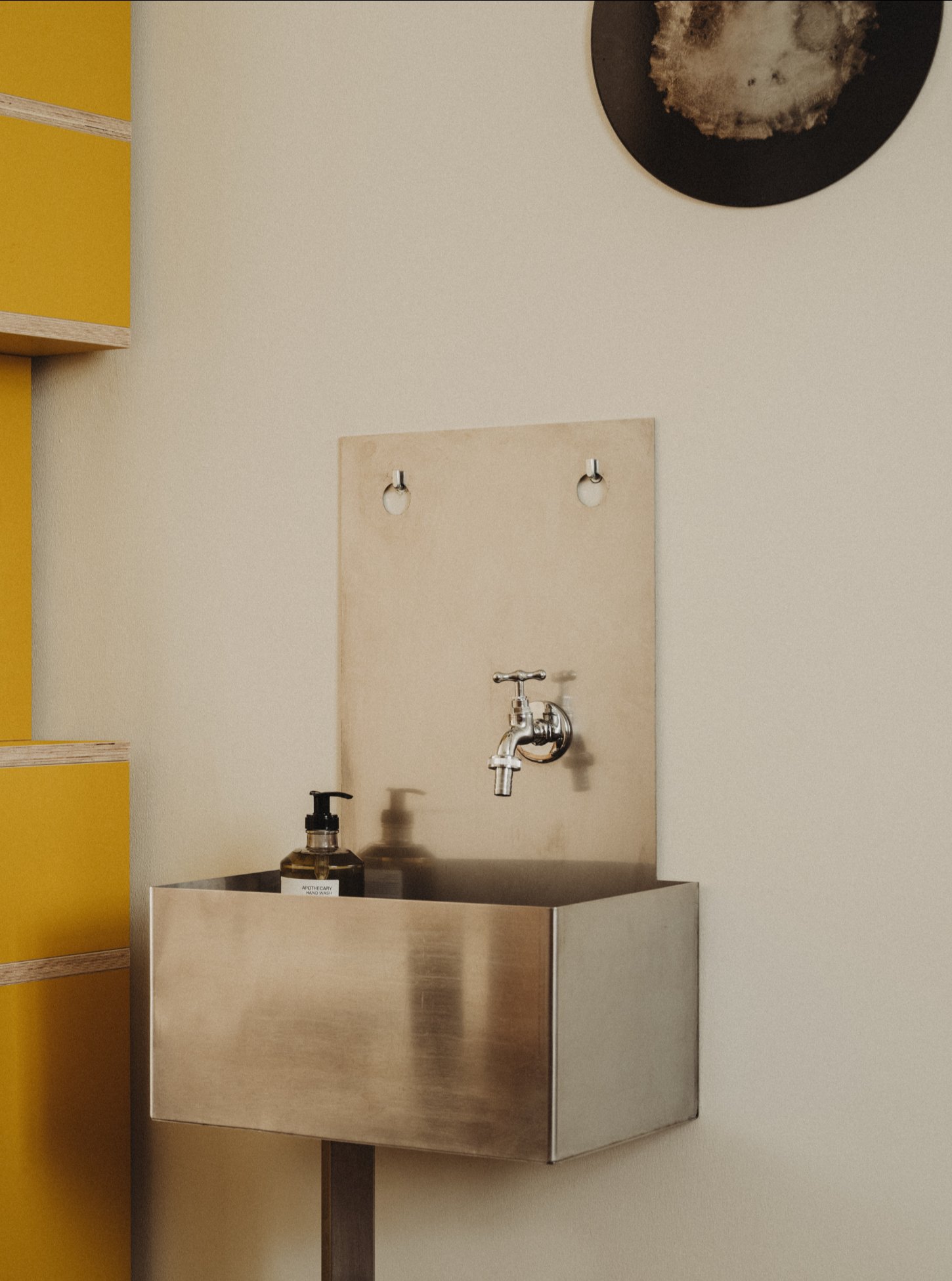 Amator Restaurant by wiercinski-studio — A wall-mounted brass tap with cross handles projects from a plywood backing panel above a stainless-steel sink basin.