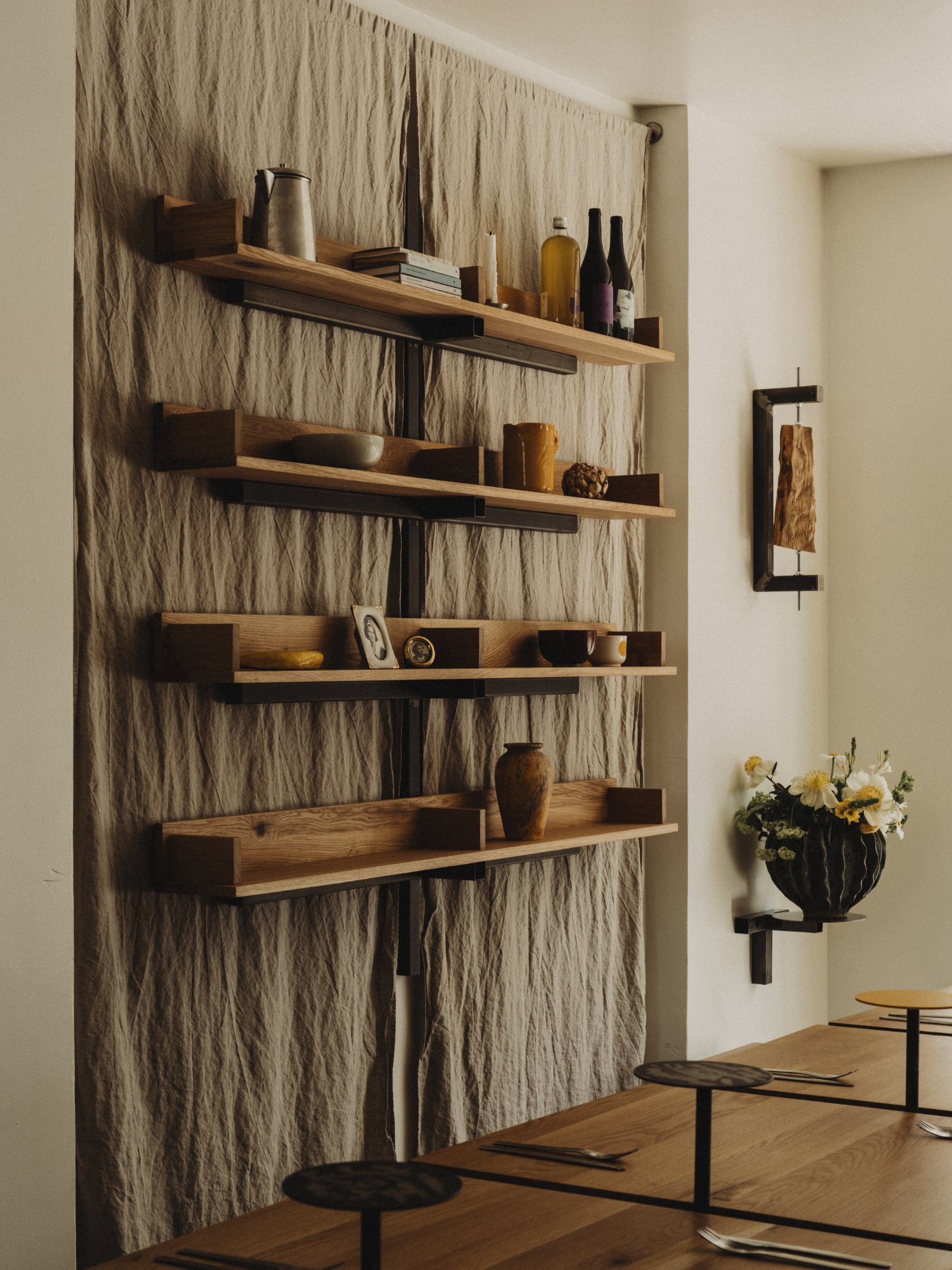 Amator Restaurant by wiercinski-studio — Four cantilevered oak shelves with light-finished tops mount horizontally against a textured linen curtain backdrop, supported by dark steel brackets.