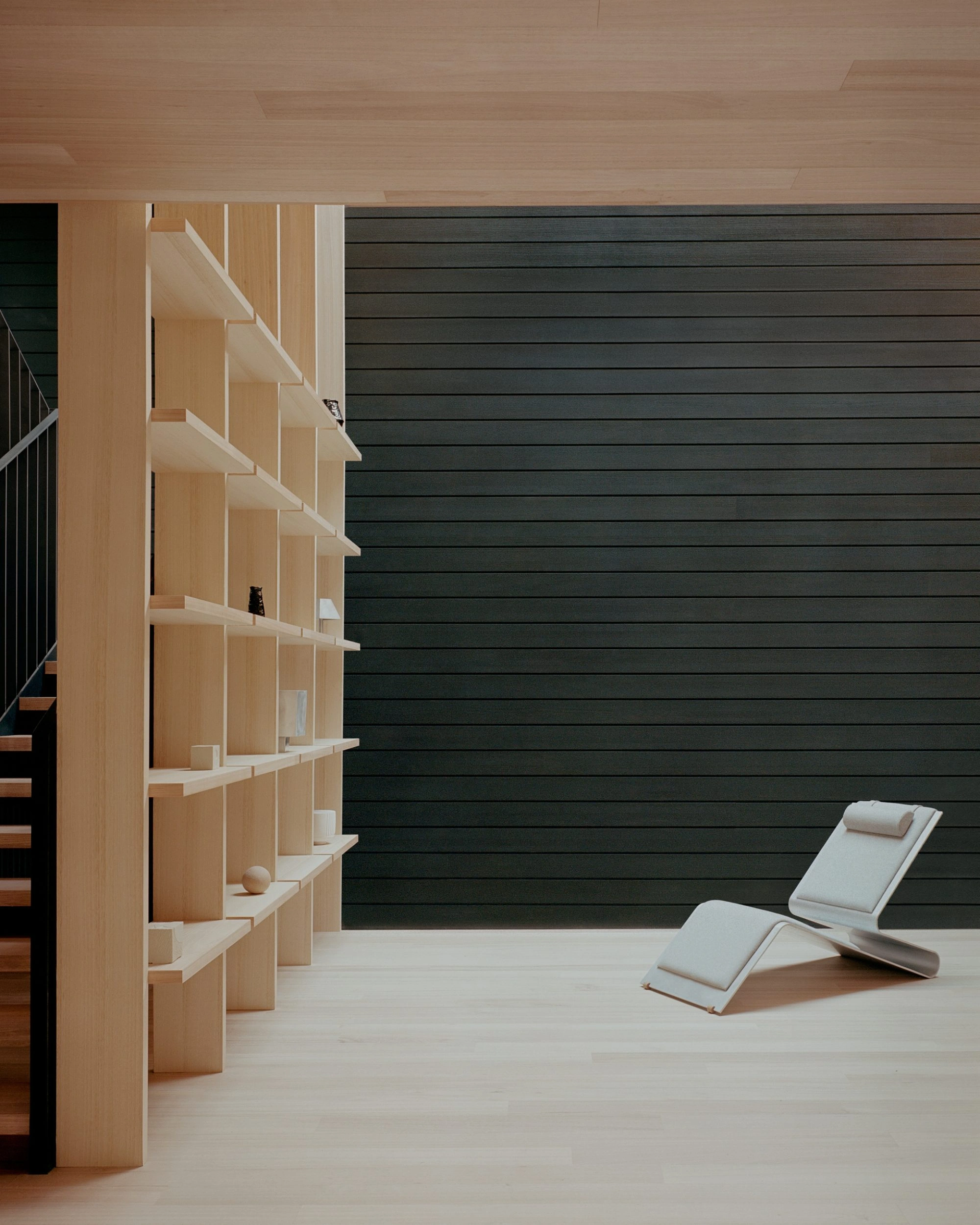 Echo House by Parabolica — A sculptural timber bookshelf in light wood dominates the left wall, its cantilevered shelves creating a rhythmic vertical composition.