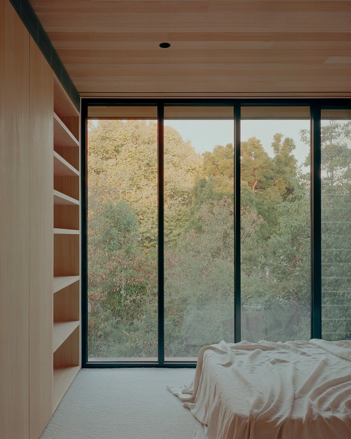Echo House by Parabolica — A bedroom corner frames dense autumn forest through floor-to-ceiling glazing with dark-framed mullions.