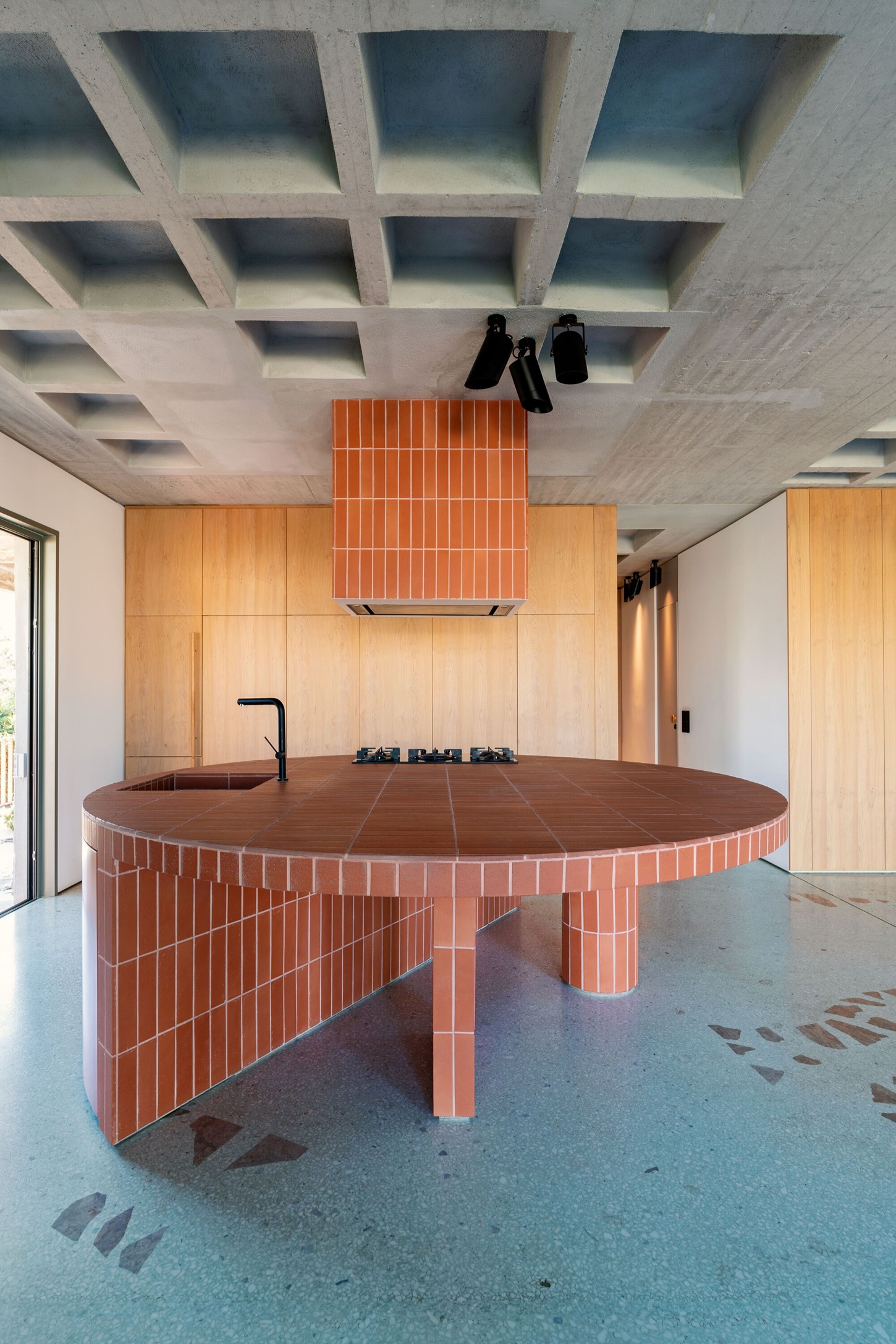 Three Object Apartment by DeMachinas + Elina Loukou — The circular kitchen island dominates the open-plan interior, its perimeter clad in vertical terracotta tile with a dark wood countertop establishing the...