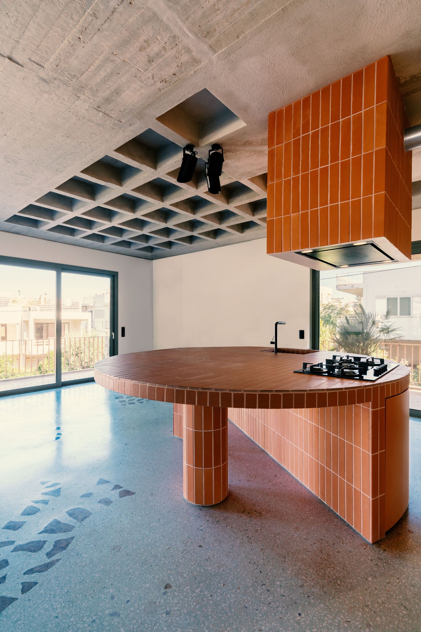 Three Object Apartment by DeMachinas + Elina Loukou — The circular kitchen island dominates the open-plan space, its perimeter wrapped in vertical terracotta tiles with a dark timber countertop.