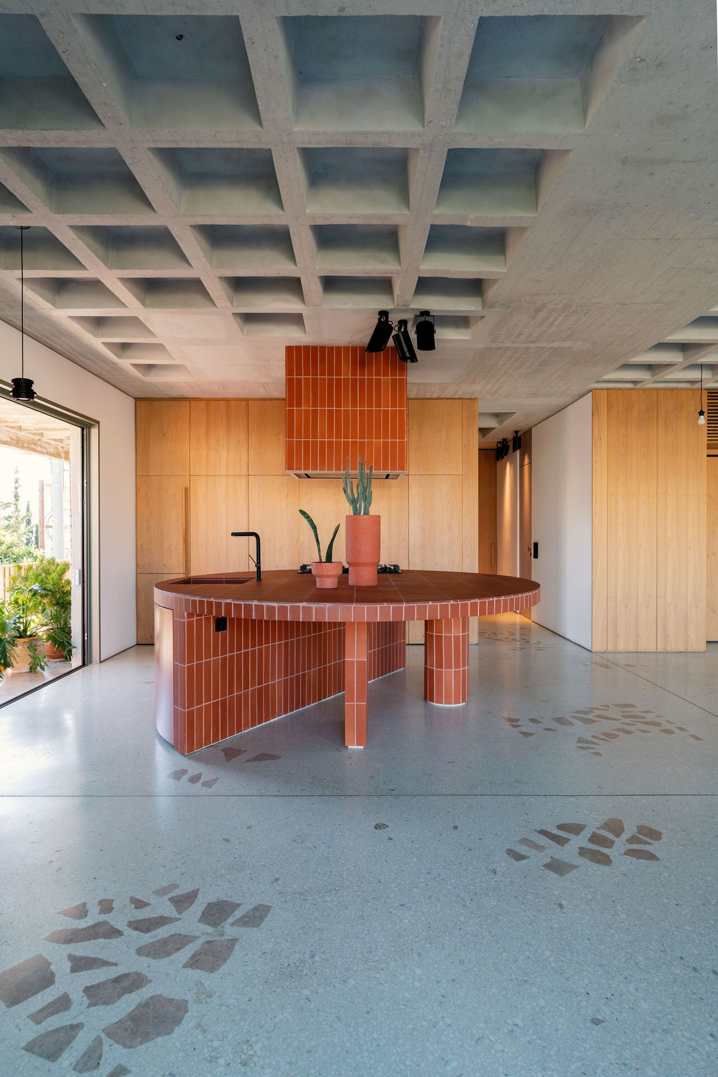 Three Object Apartment by DeMachinas + Elina Loukou — The circular kitchen island dominates the open-plan ground floor, its perimeter wrapped in vertical terracotta tile with a dark wood countertop that anchors...