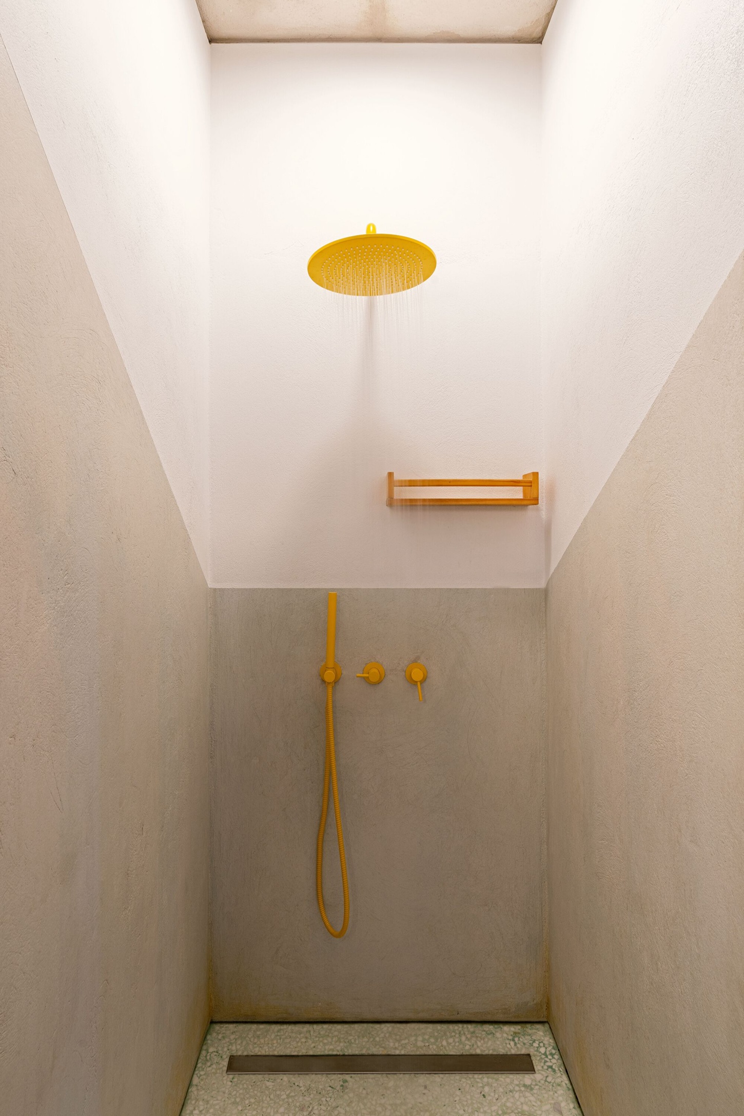 Three Object Apartment by DeMachinas + Elina Loukou — A shower enclosure defined by minimalist fixtures in golden-yellow metal against rendered concrete walls.