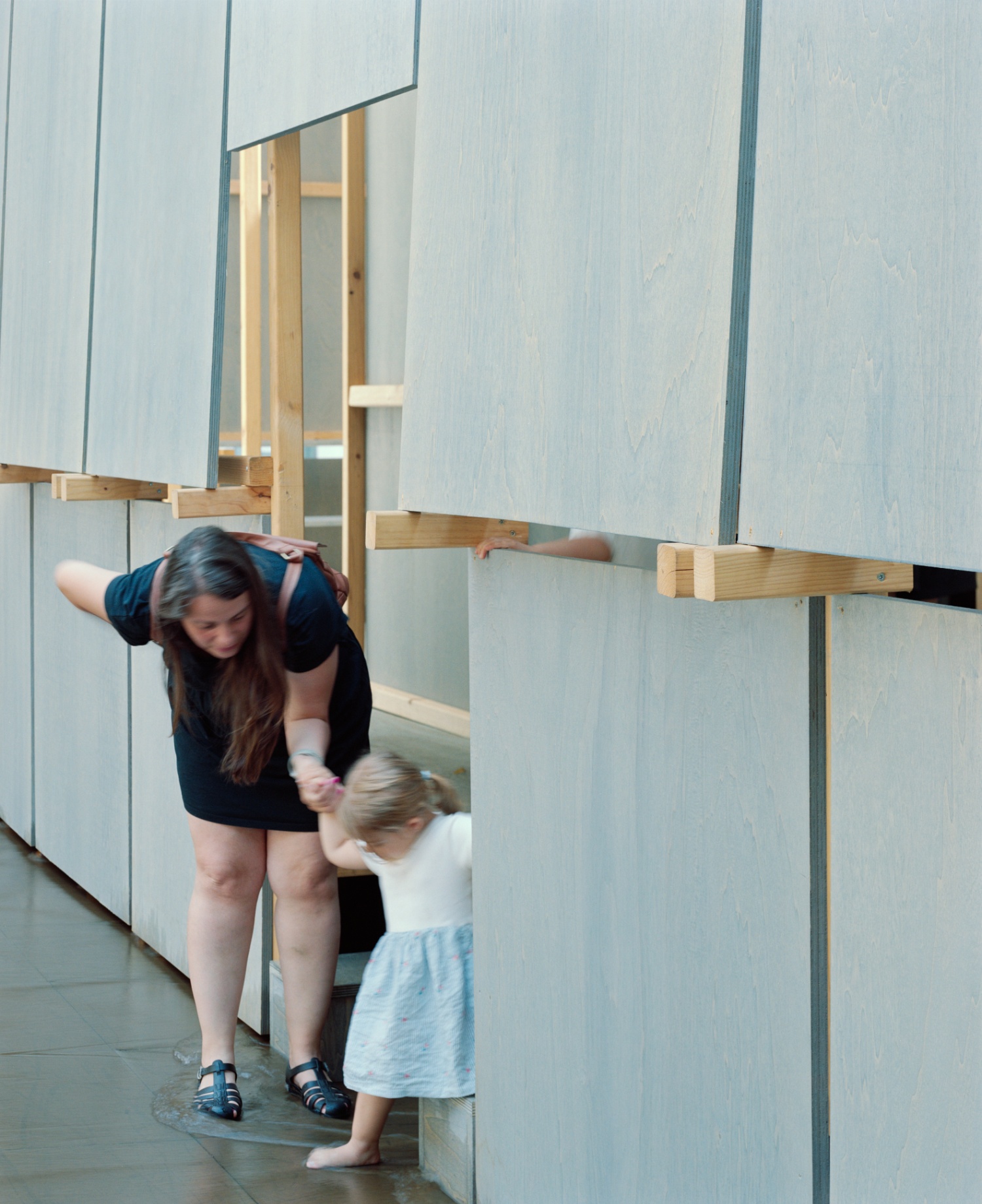 Round About Baths by Leopold Banchini — An adult and child navigate the timber-framed threshold of the temporary bathhouse, their figures backlit against the recessed dark doorway.