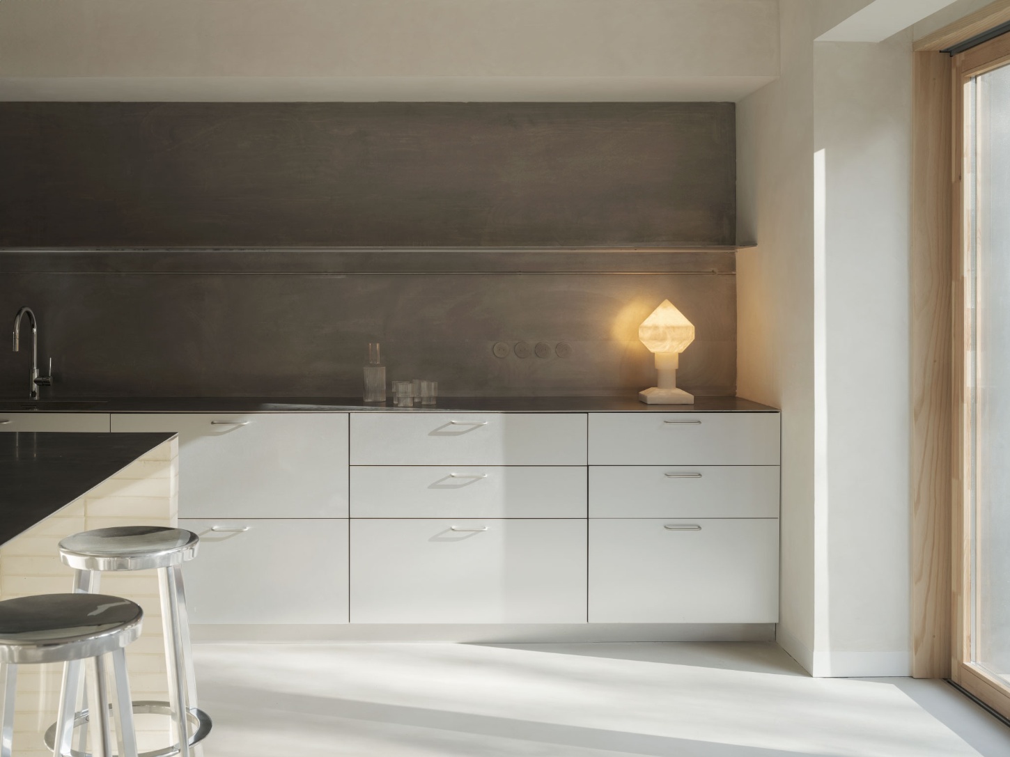 Casa SO by Studio DIIR — A modernist kitchen bathed in south-facing natural light, where white lacquered cabinetry and a charcoal-rendered backsplash establish a studied chromatic...