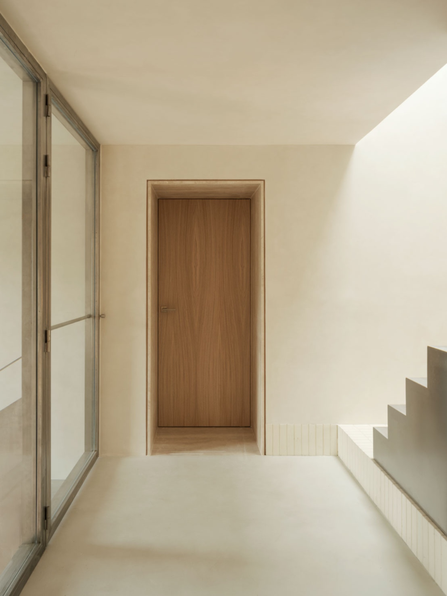 Casa SO by Studio DIIR — A timber-framed doorway anchors the end of a narrow corridor, its warm wood grain contrasting sharply with cream plaster walls.