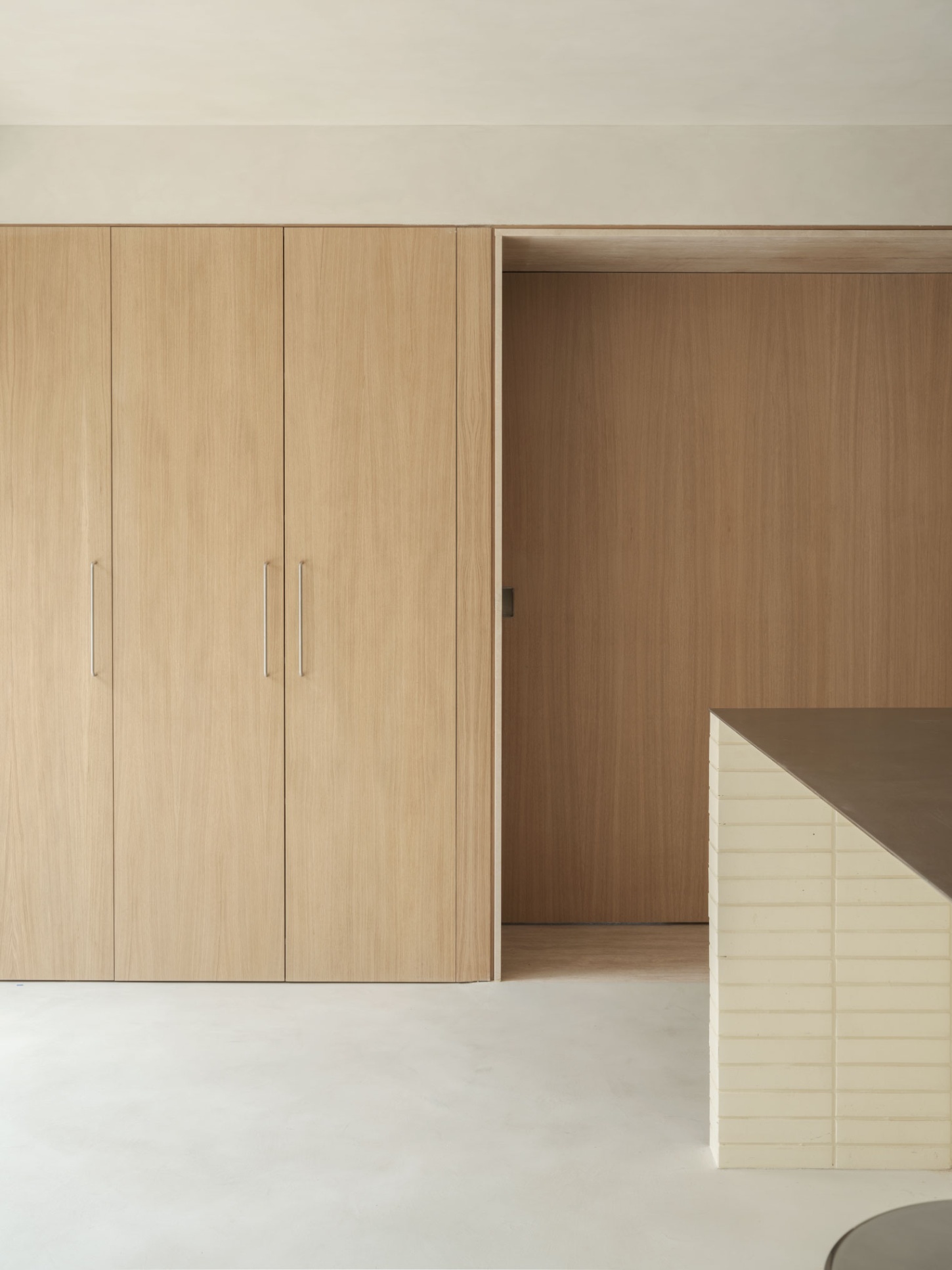 Casa SO by Studio DIIR — A timber-lined passage frames the transition between two domestic zones, with light-oak cabinetry dominating the left wall, its vertical grain articulated...