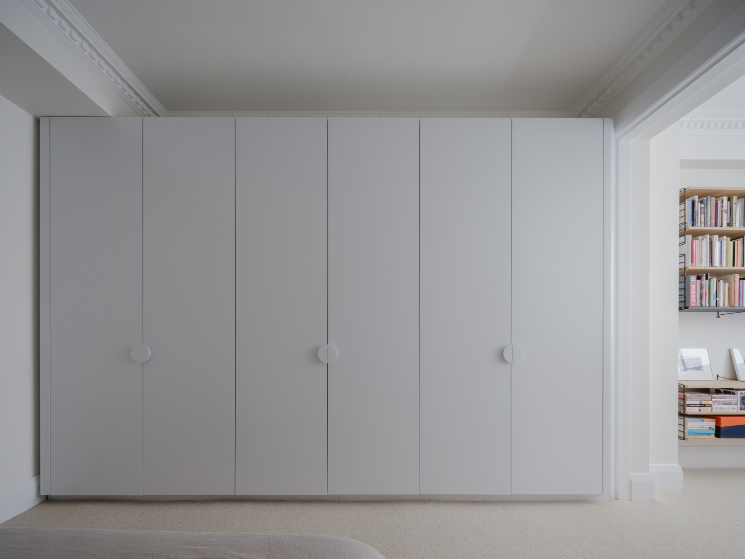 Onslow Gardens Apartment by Youssofzay Hart — A wall of built-in cabinetry dominates the bedroom interior, its six vertical panel doors rendered in soft grey-white with minimal circular handles creating...