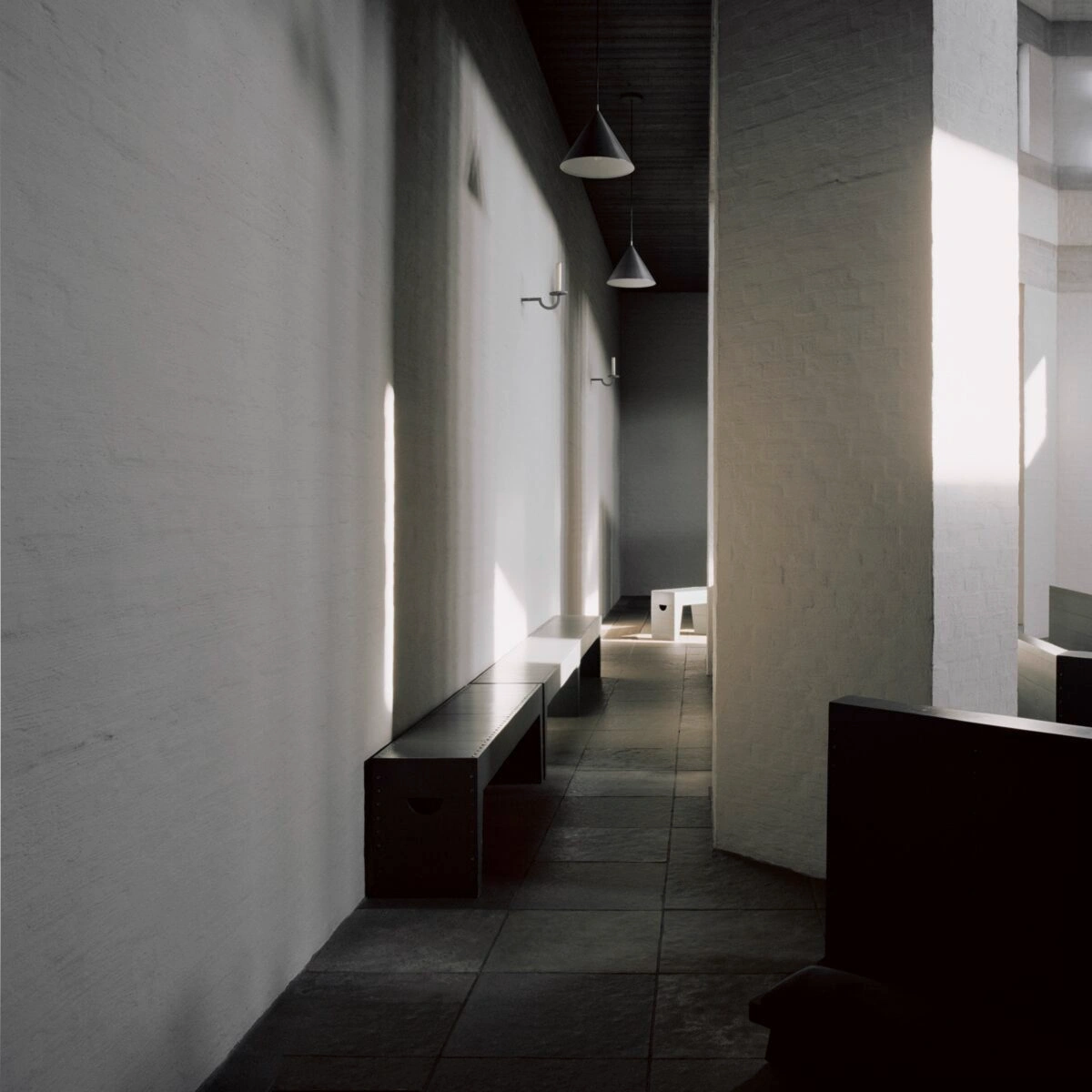 Roosenberg Abbey by Dom Hans van der Laan — A narrow interior corridor defined by offset white plaster walls and dark timber benches positioned asymmetrically along the floor edge.
