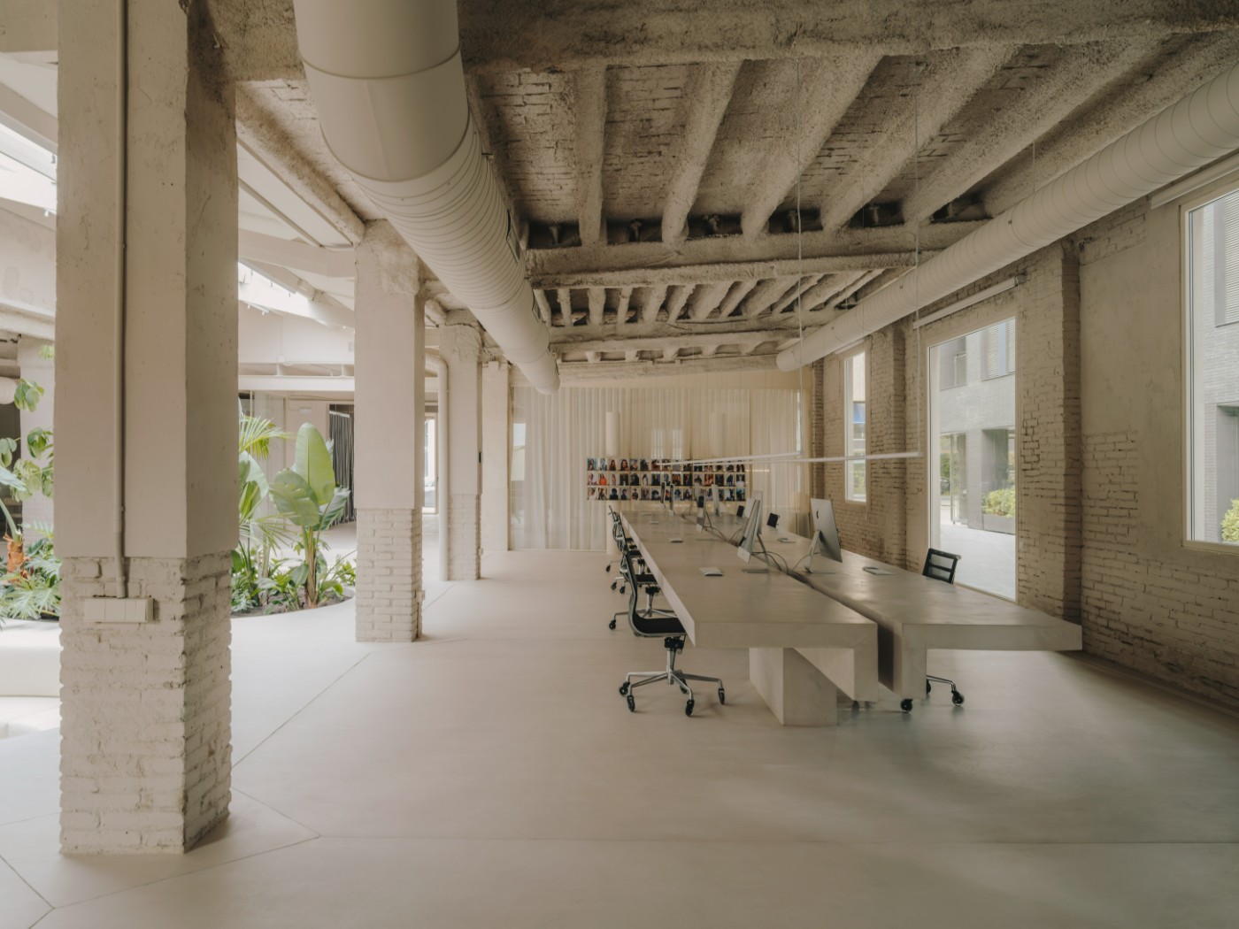 Blow Studio by Isern Serra — A linear workspace stretches beneath exposed concrete beams and a central skylight, anchored by a continuous curved bench upholstered in cream fabric that...