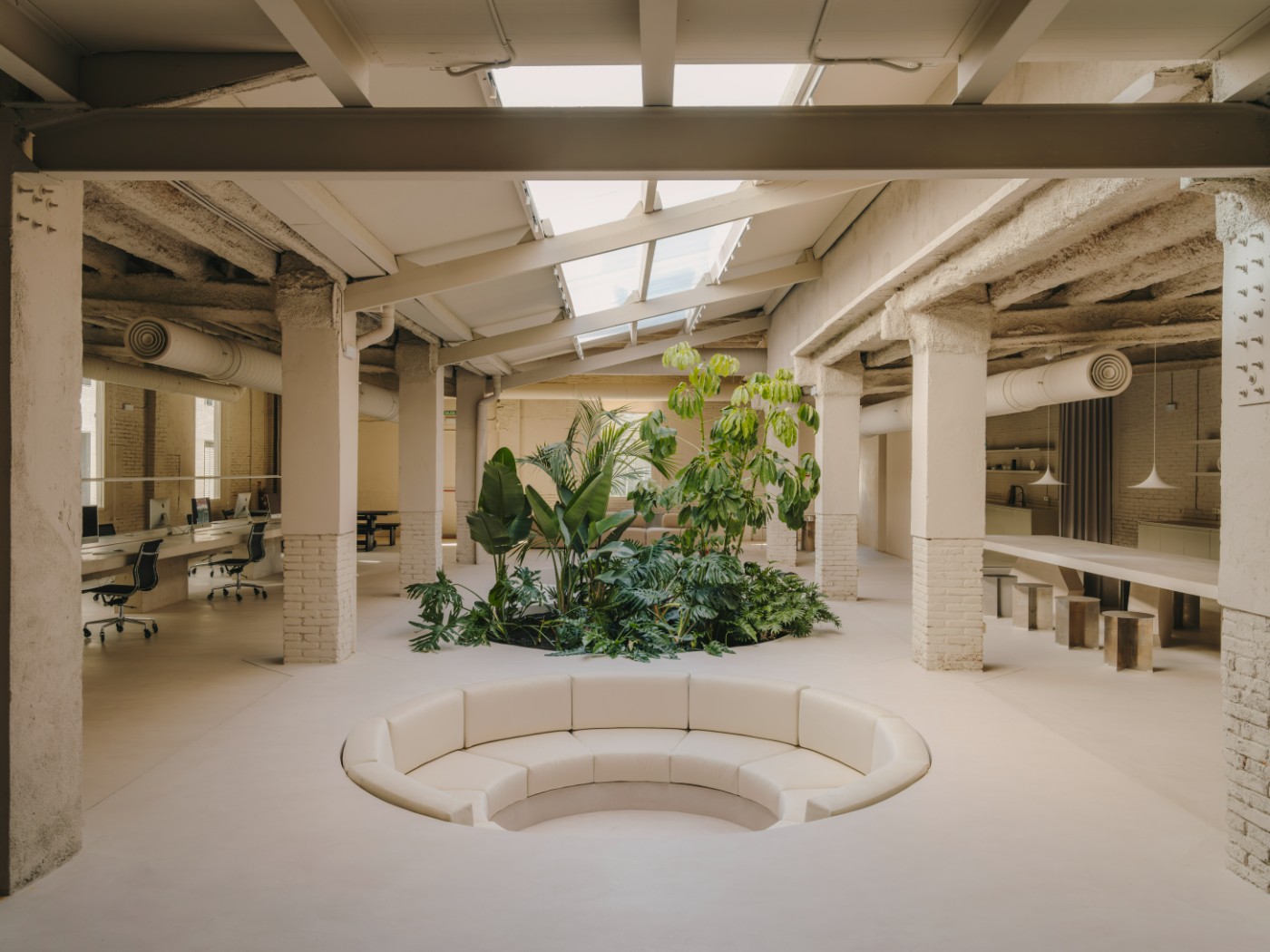 Blow Studio by Isern Serra — A centralized recessed seating pit with curved cream-upholstered bench segments anchors the ground plane of this converted warehouse interior.