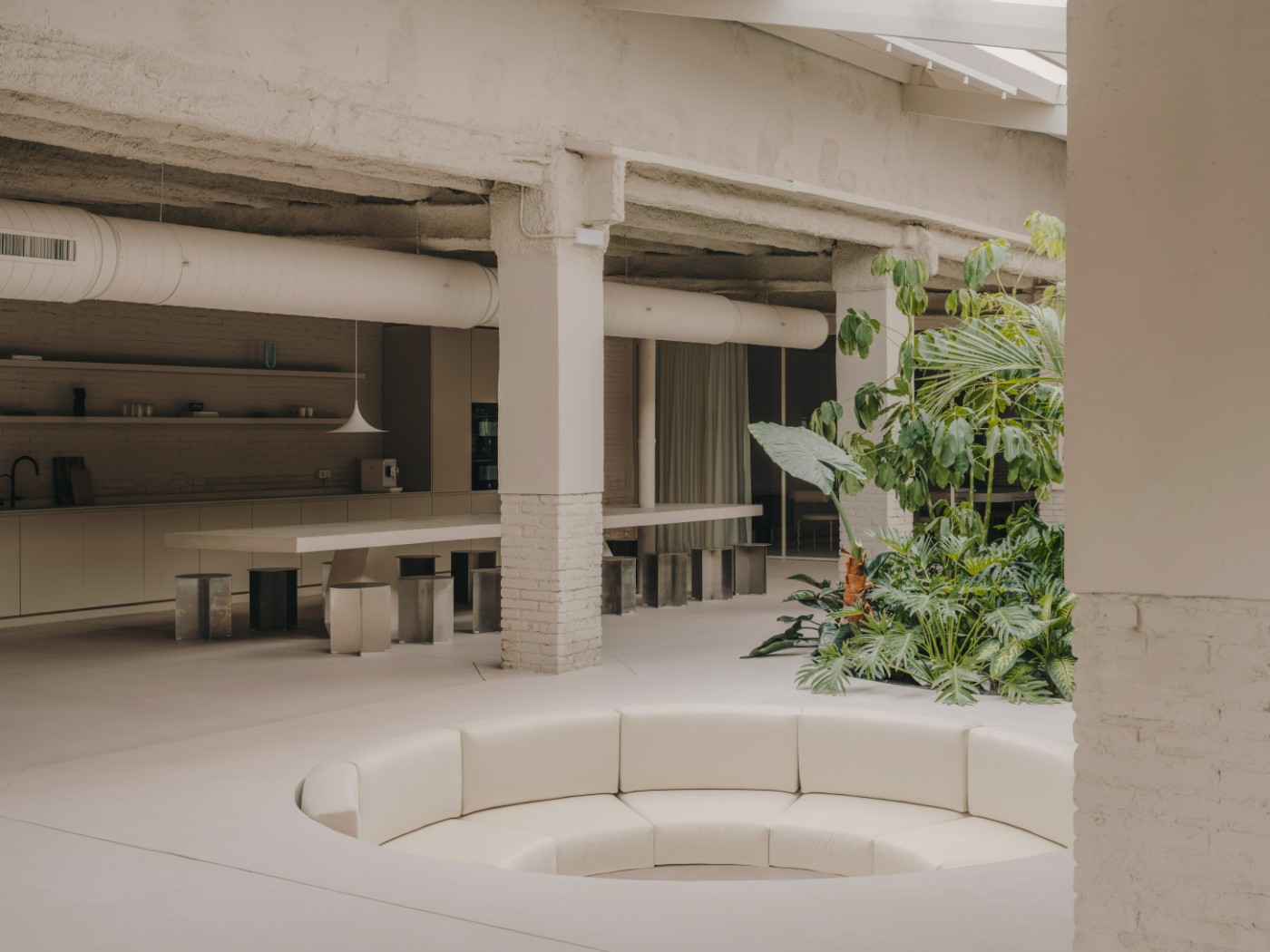 Blow Studio by Isern Serra — A recessed circular seating pit anchors the central atrium, its curved cream-colored upholstered bench forming a continuous ring within the polished...