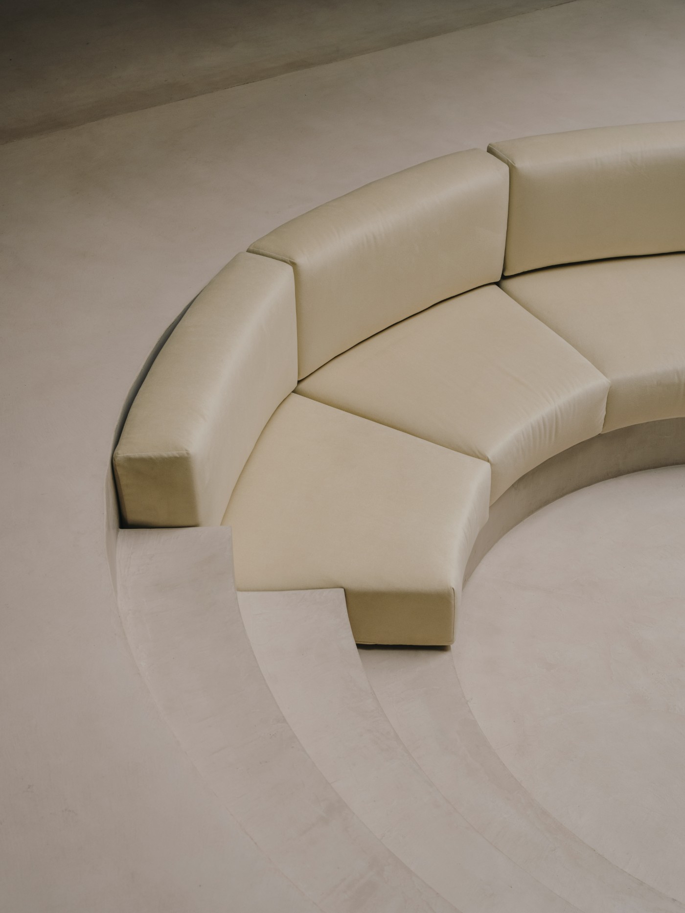 Blow Studio by Isern Serra — A curved cream-colored upholstered bench forms a continuous seating surface recessed into the polished concrete floor, its modular sections creating an...