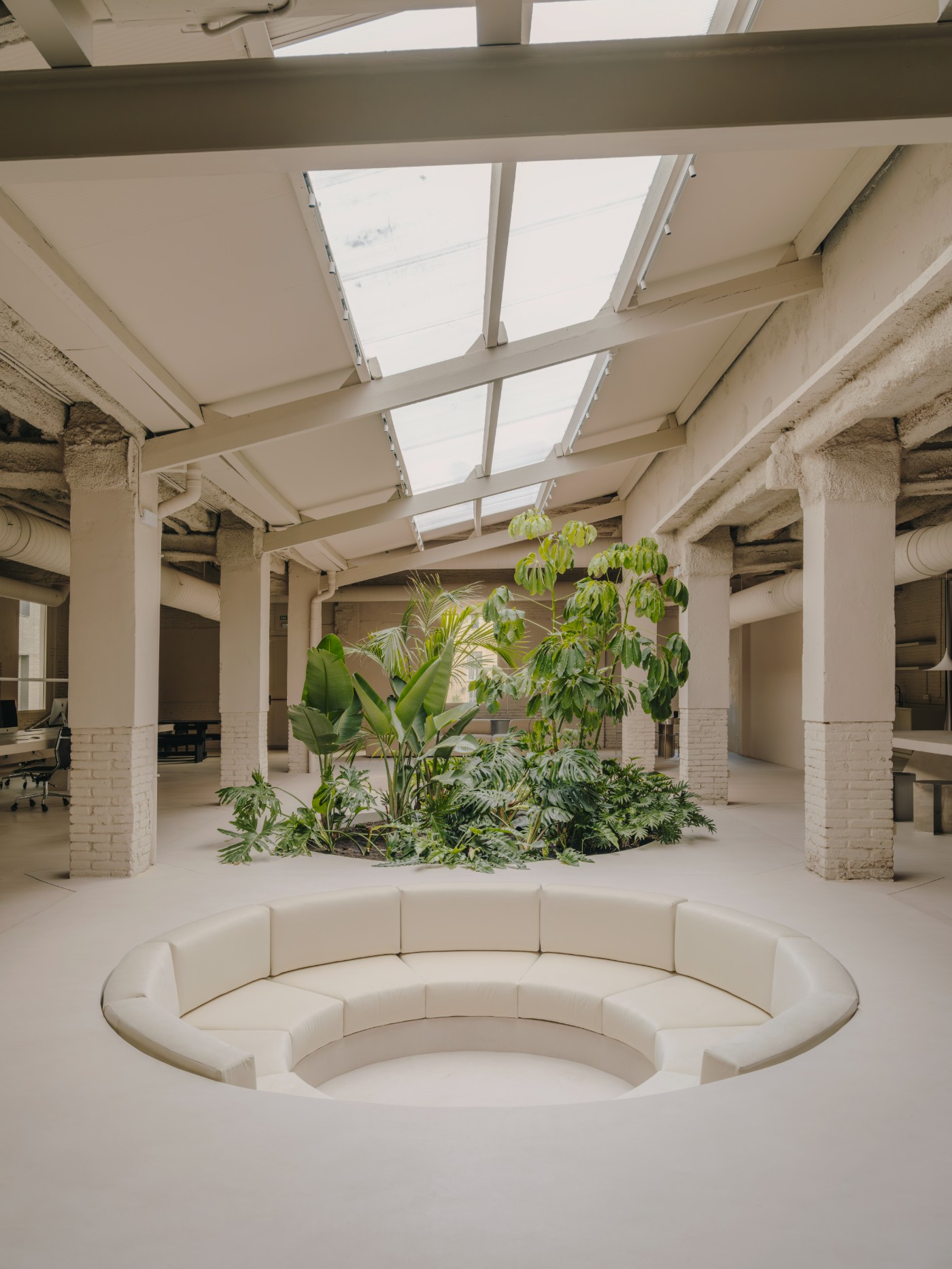 Blow Studio by Isern Serra — A recessed circular seating pit anchors the central volume of this converted warehouse, its cream-colored upholstered bench forming a continuous curved...