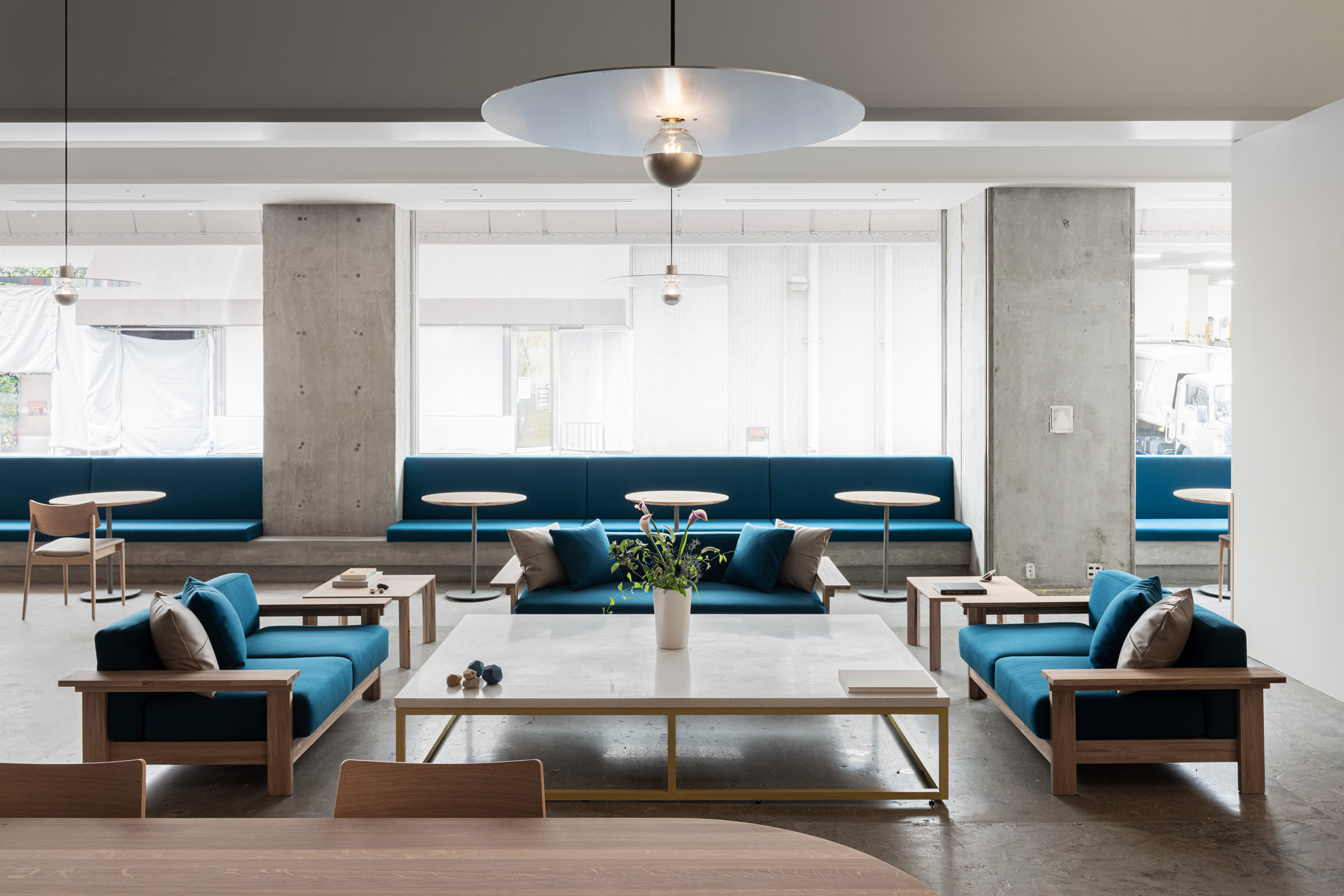 Blue Bottle Coffee Kobe Hankyu Cafe by Keiji Ashizawa — The cafe's lounge seating demonstrates a symmetrical arrangement of teal upholstered low seating with timber frames, flanking a white marble-topped coffee...