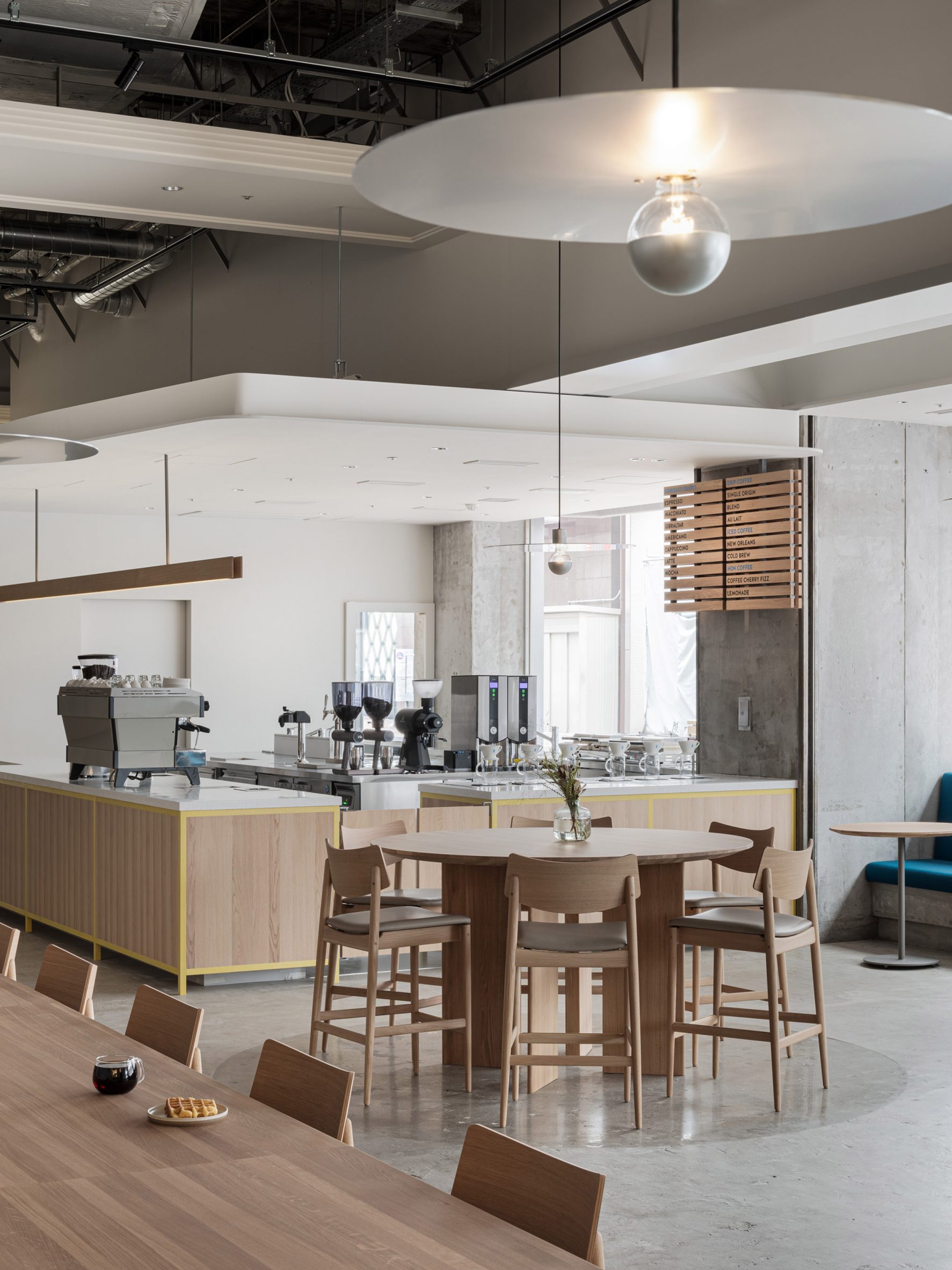 Blue Bottle Coffee Kobe Hankyu Cafe by Keiji Ashizawa — A circular timber counter anchors the cafe interior, surrounded by wooden chairs with woven seats.