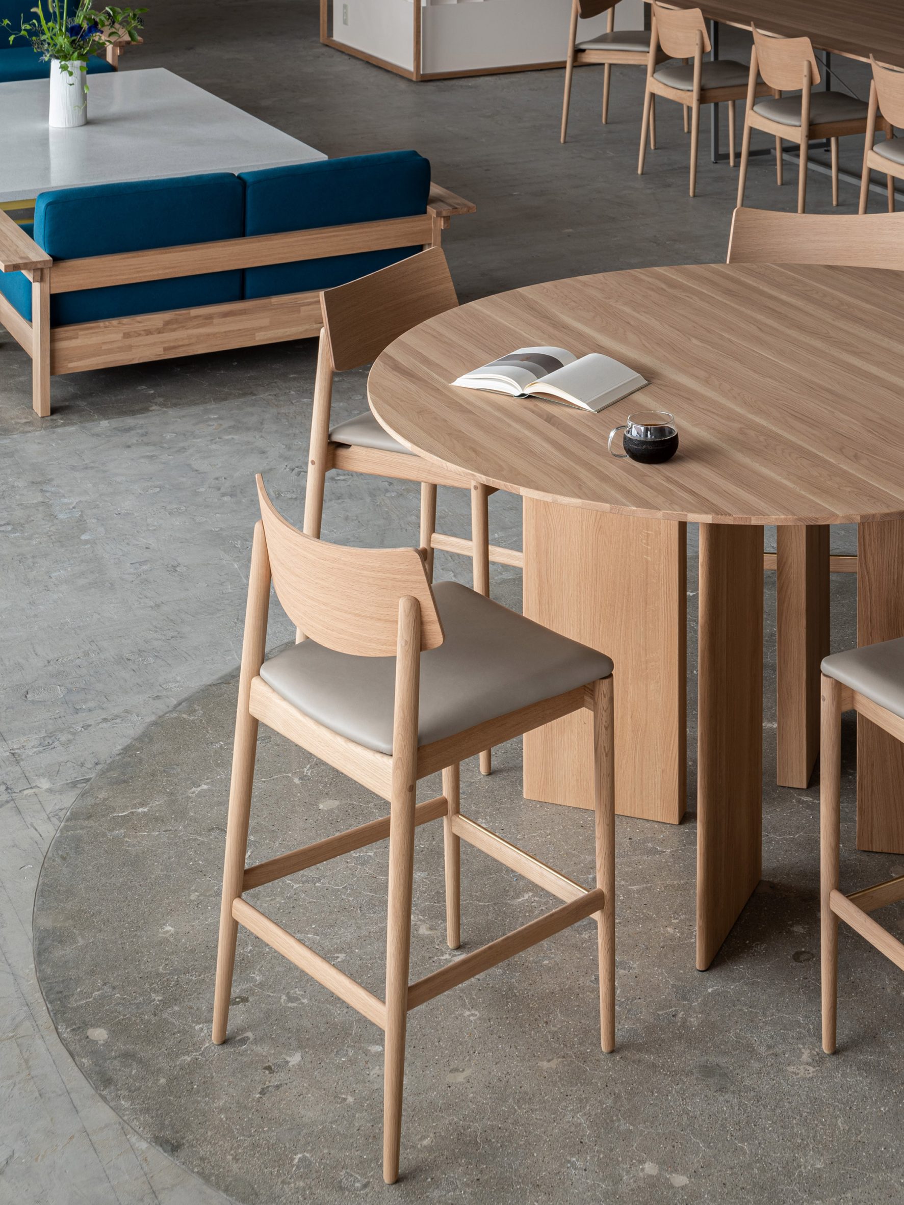 Blue Bottle Coffee Kobe Hankyu Cafe by Keiji Ashizawa — A round oak table with vertical grain detailing anchors the cafe seating area, its curved geometry contrasting with the rectilinear forms dominating the space.