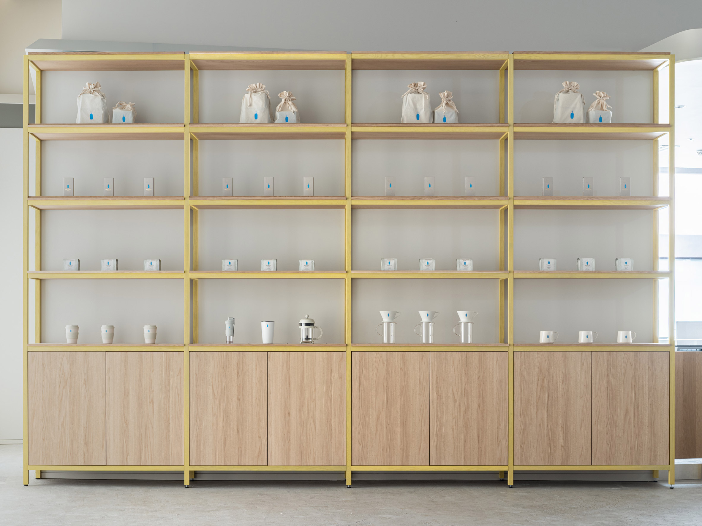 Blue Bottle Coffee Kobe Hankyu Cafe by Keiji Ashizawa — A modular shelving system with brass-toned metal framing organizes the retail display across four vertical bays.