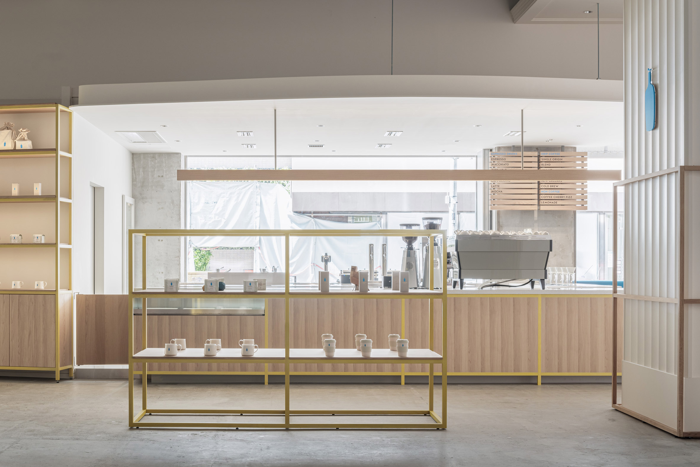 Blue Bottle Coffee Kobe Hankyu Cafe by Keiji Ashizawa — The counter zone at Blue Bottle Coffee Kobe Hankyu Cafe demonstrates a layered display strategy through brass-framed glass vitrines positioned at varying...