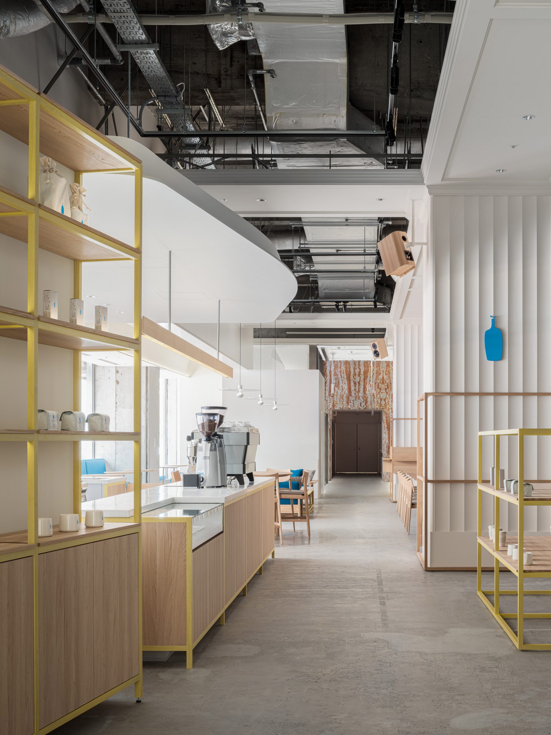 Blue Bottle Coffee Kobe Hankyu Cafe by Keiji Ashizawa — The café's 173-square-metre corridor stretches linearly between yellow-framed timber shelving units and a timber-clad service counter.