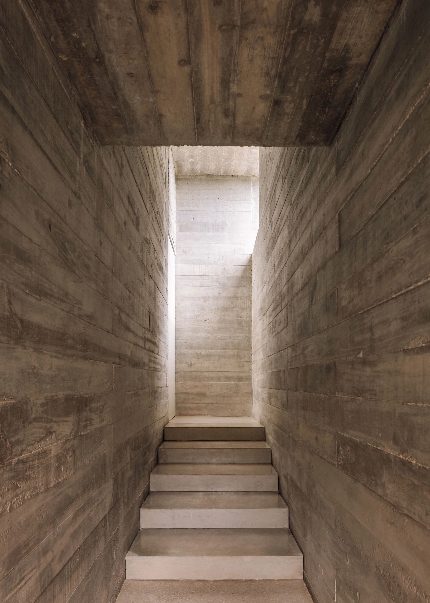 Frame House by Pedro Domingos Arquitectos — A linear concrete staircase ascends through a narrow passage formed by two weathered board-formed concrete walls.
