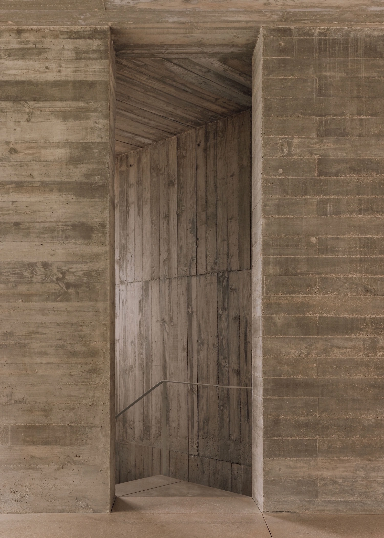 Frame House by Pedro Domingos Arquitectos — A narrow timber-lined passage recedes into shadow, its board-formed concrete walls framing a vertical void.