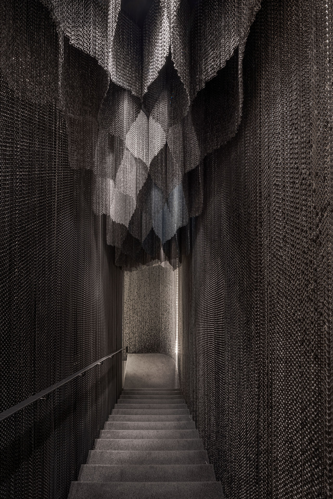 Casa Batlló Stairs & Atrium by Kengo Kuma — A narrow stairwell descends into shadow, flanked by floor-to-ceiling cascading aluminum chain curtains that fragment incoming light into shifting patterns...