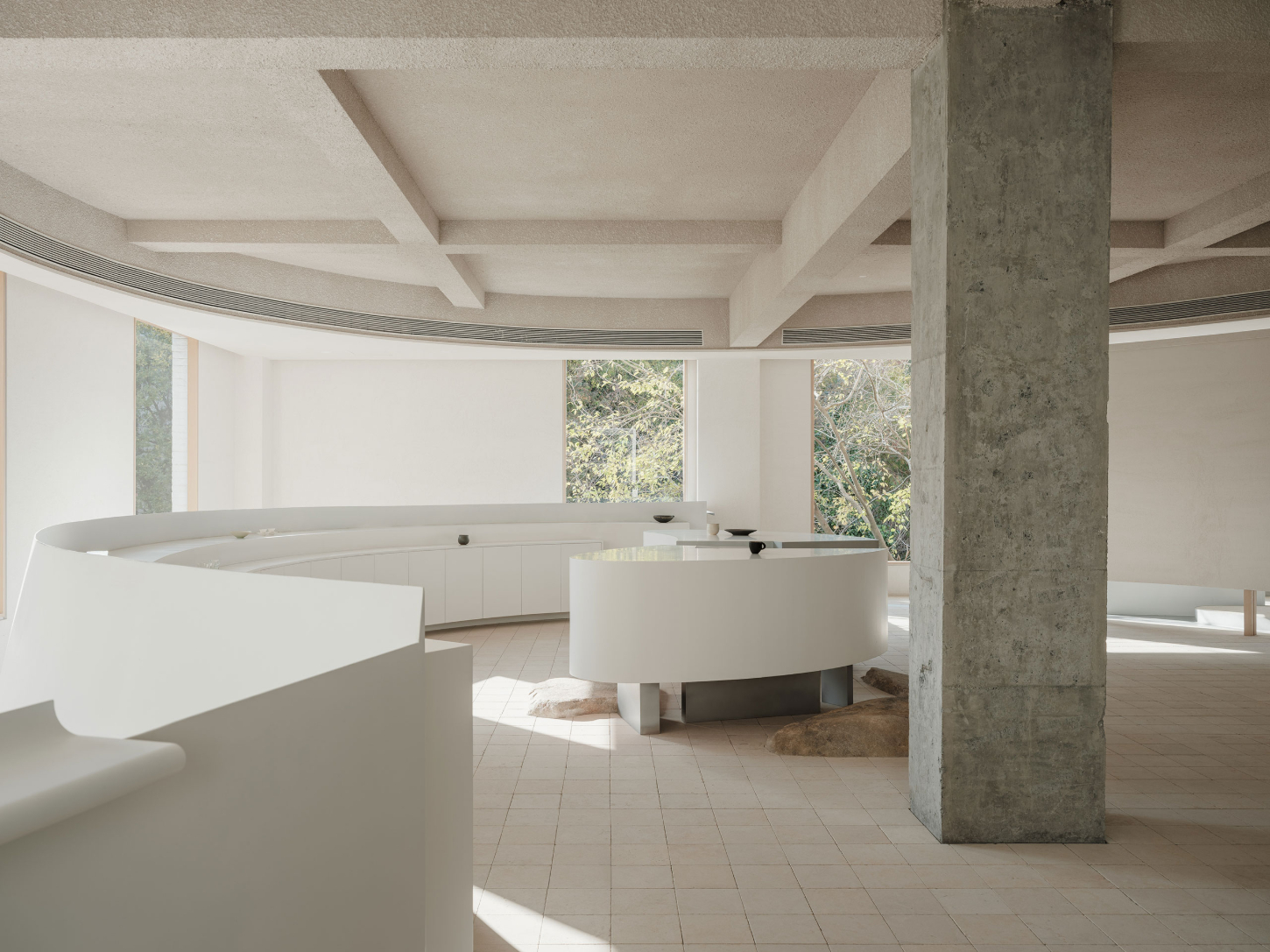 JIA Garden by Mountain Soil — An interior showroom featuring two elongated white display tables with organic, leaf-shaped profiles positioned asymmetrically within a coffered concrete...