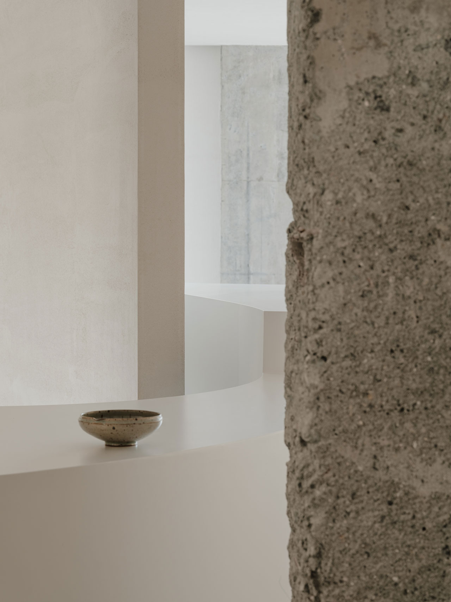 JIA Garden by Mountain Soil — A minimalist display interior featuring a white curved table with organic edges set against raw concrete and rammed-earth walls.