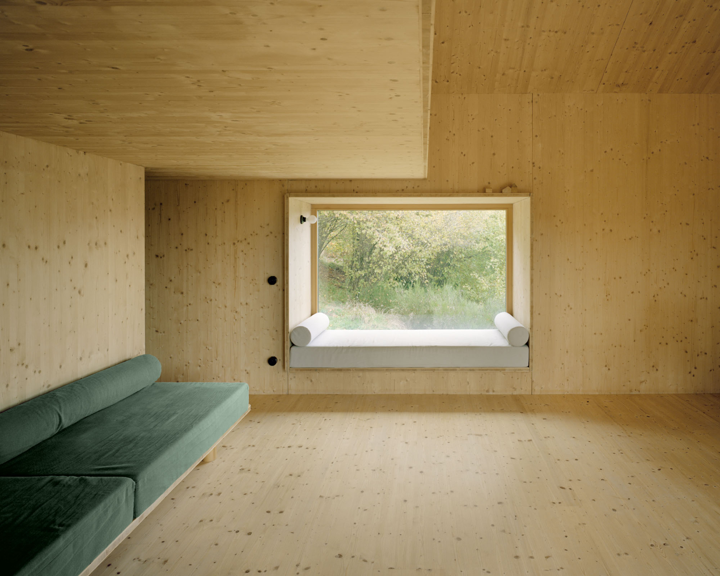 Maison Hiltiti captured by Rasmus Norlander by Atelier Ordinaire — A bedroom interior clad entirely in cross-laminated timber (CLT), featuring a built-in window seat with white upholstery positioned to frame views of leafy...