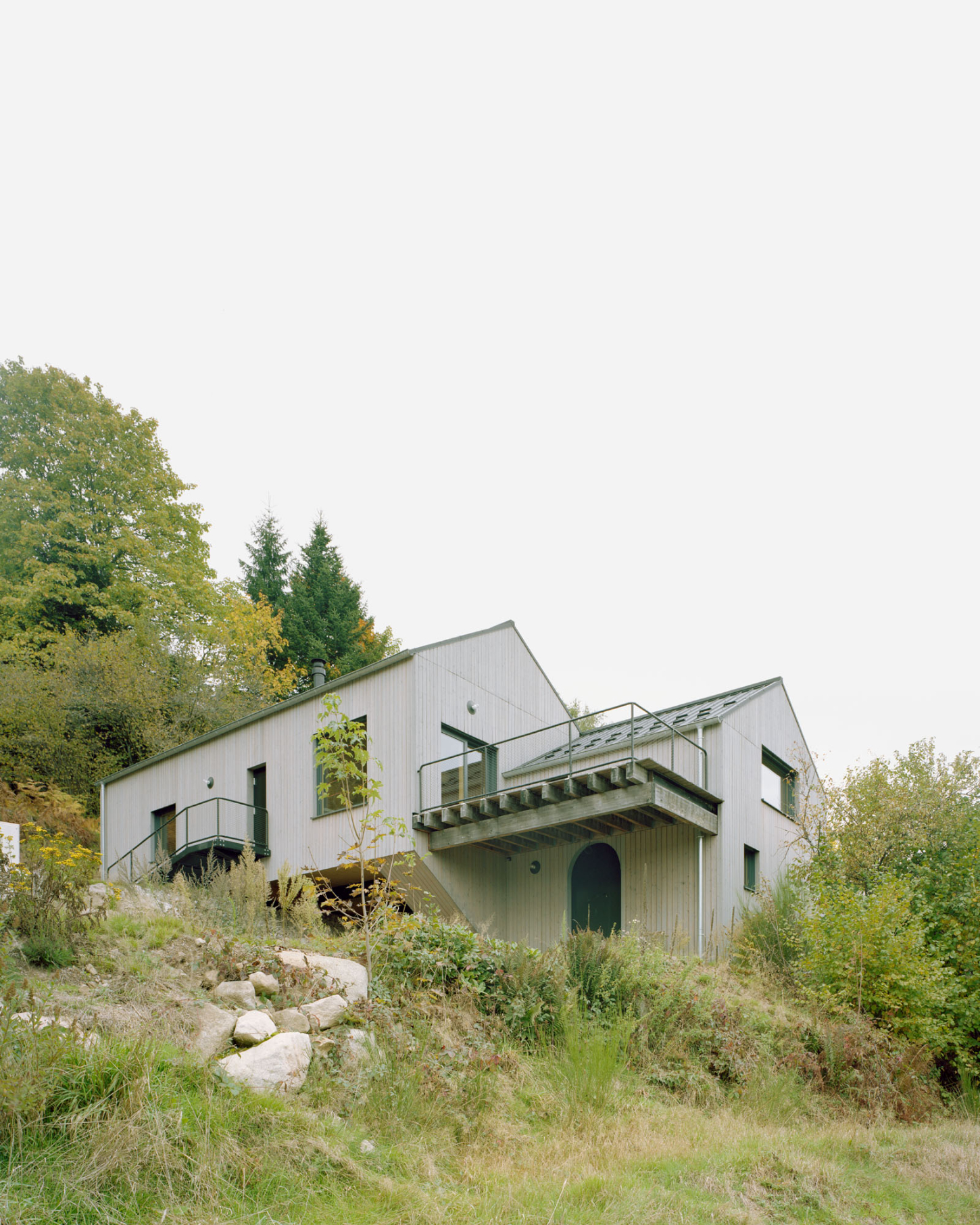 Maison Hiltiti captured by Rasmus Norlander by Atelier Ordinaire — Maison Hiltiti sits embedded within a sloping landscape of wild grasses and boulders, its timber-clad volumes ascending the hillside in staggered configuration.