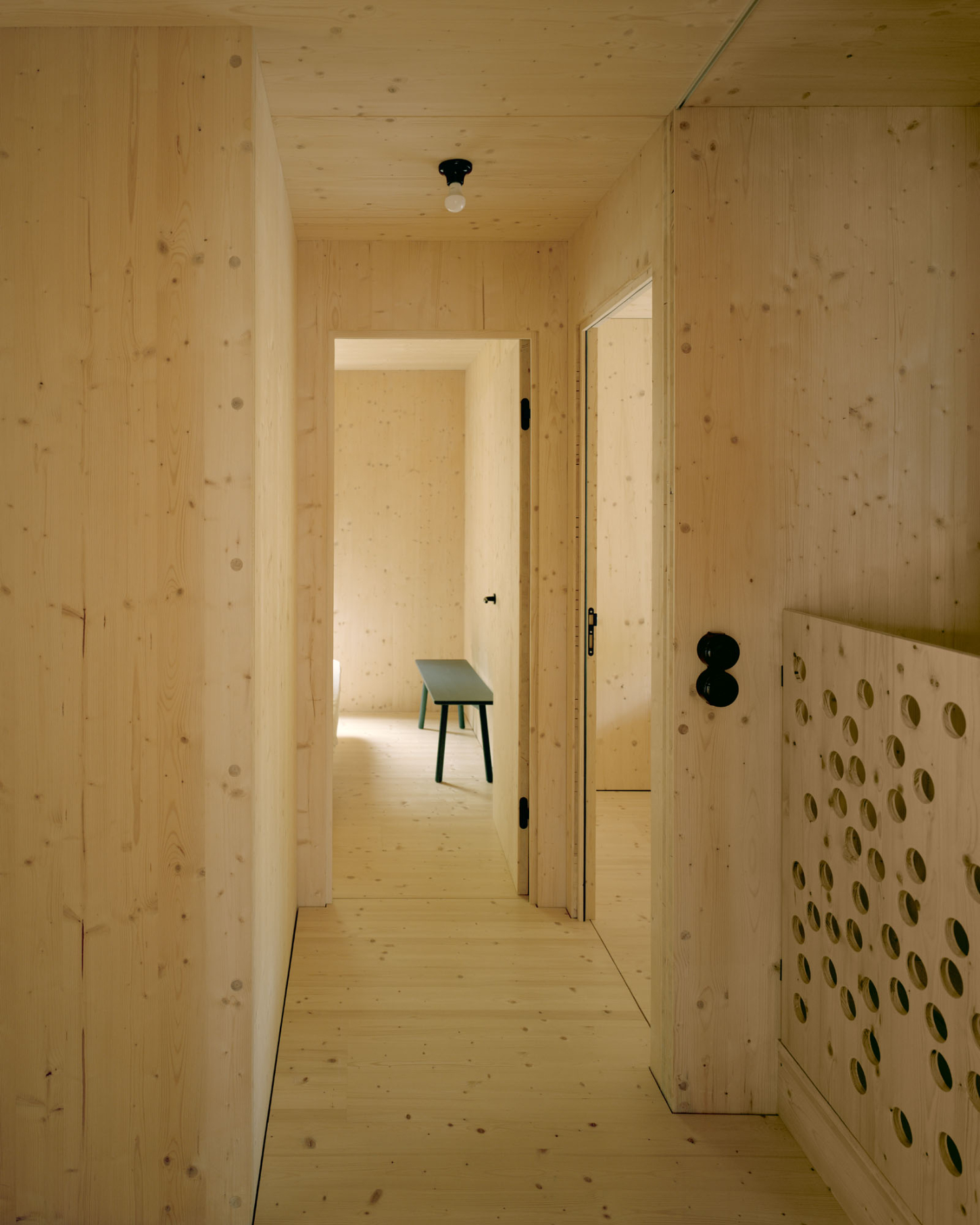 Maison Hiltiti captured by Rasmus Norlander by Atelier Ordinaire — A narrow corridor lined entirely in cross-laminated timber extends toward distant rooms, its continuous wood flooring and wall planes creating a linear...