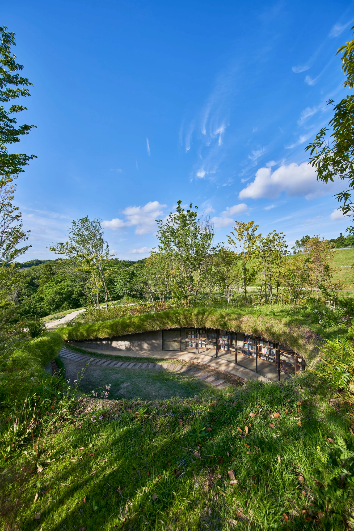 Library in the Earth by Hiroshi Nakamura & NAP — A recessed library entrance set into an earthen berm, accessed via a stepping stone pathway that recedes toward a dark timber-framed portal.