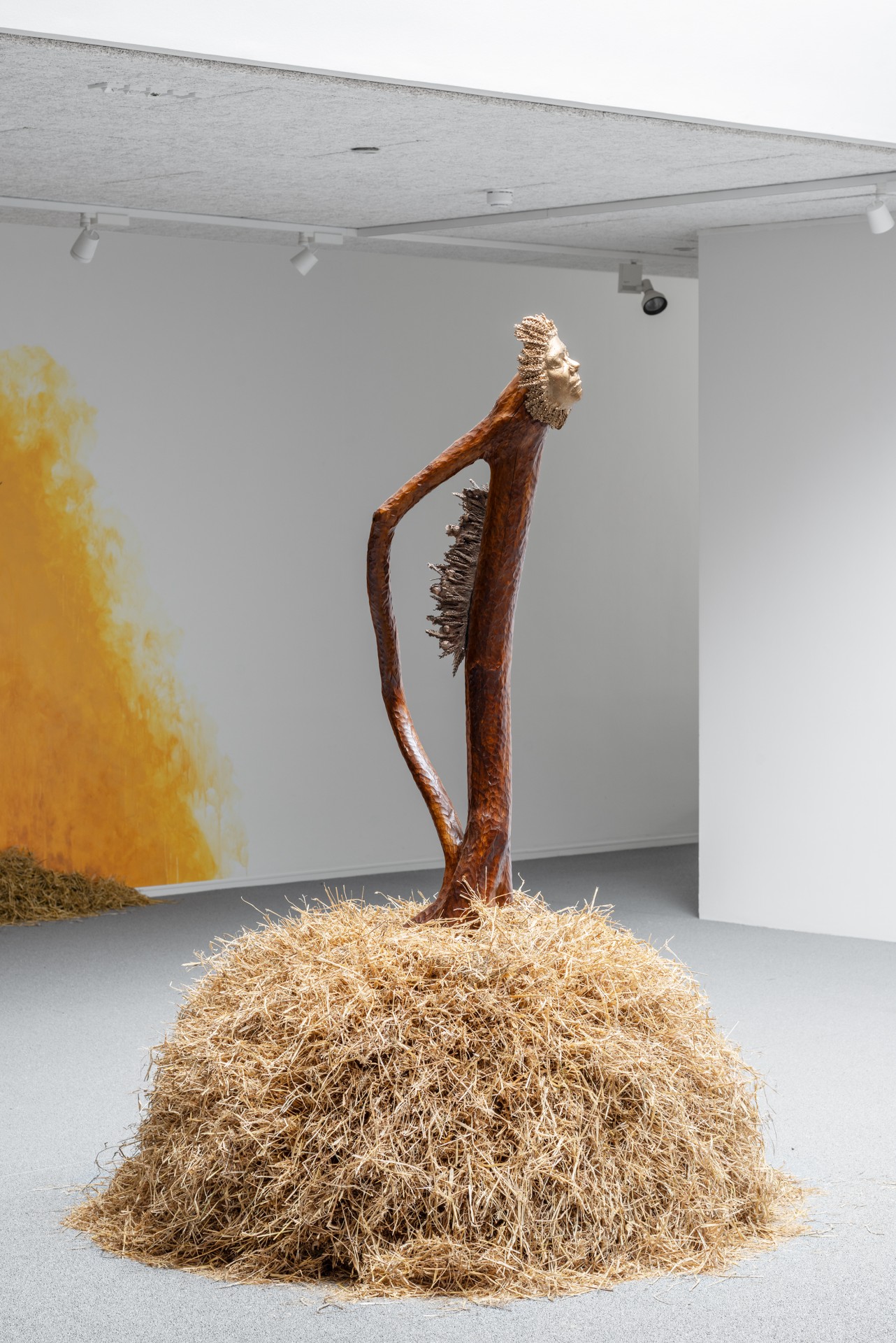 Bleak Harvest by Anna Bak at Vestjyllands Kunstpavillon — A wooden sculptural figure emerges from a dense mound of loose straw, its elongated body curved in a contorted posture.