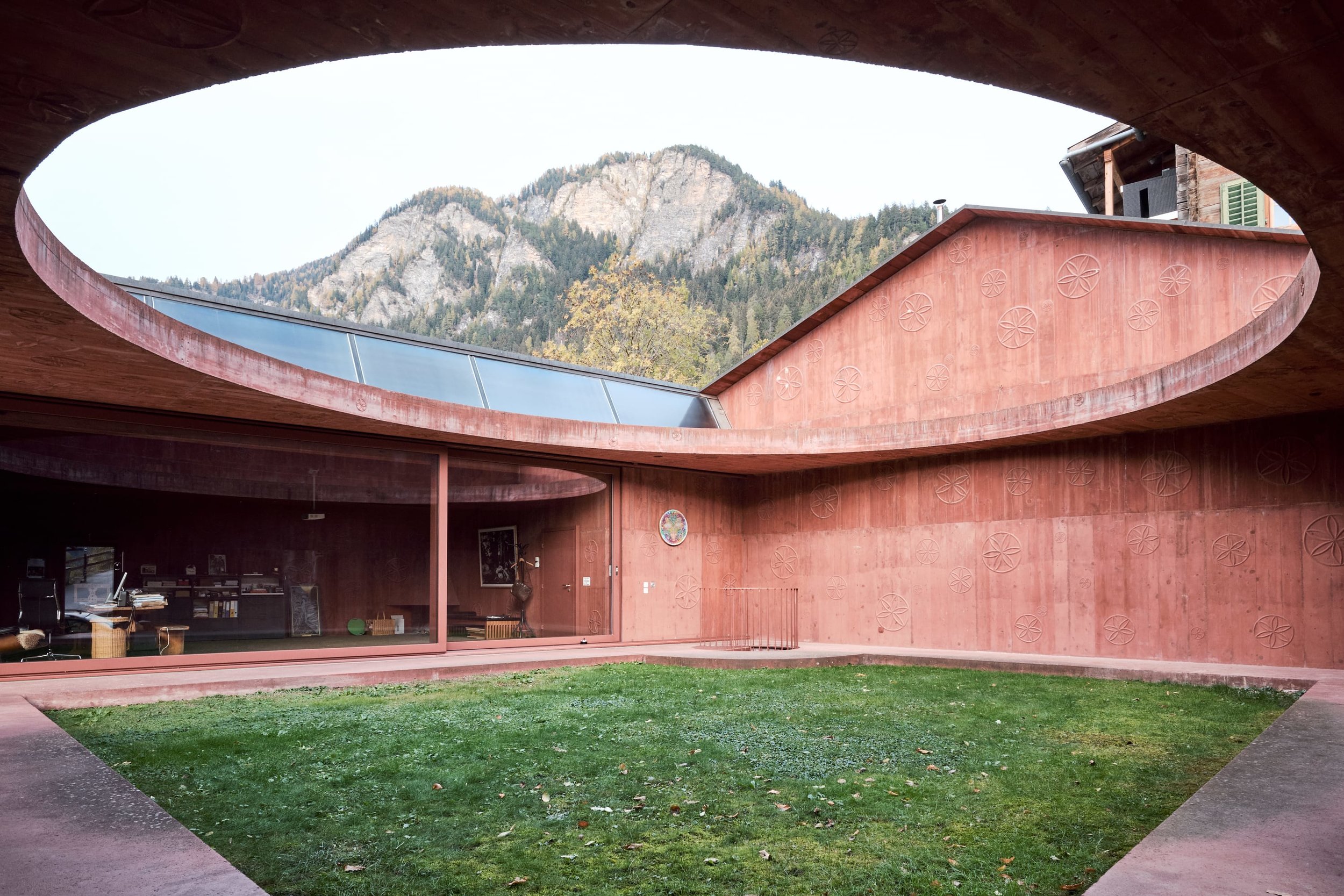 Atelier Bardill by Valerio Olgiati — A monolithic curved concrete structure wraps around a grass courtyard, its terracotta-rendered facade punctuated by an embossed geometric pattern of...