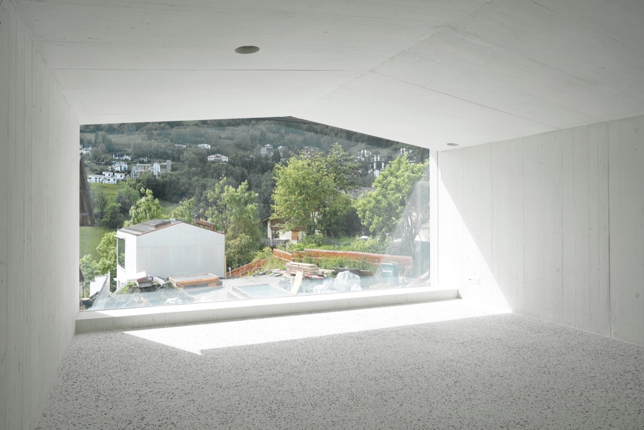 House in Laax by Valerio Olgiati — A rendered white interior wall frames an expansive horizontal window opening onto a hillside village landscape.