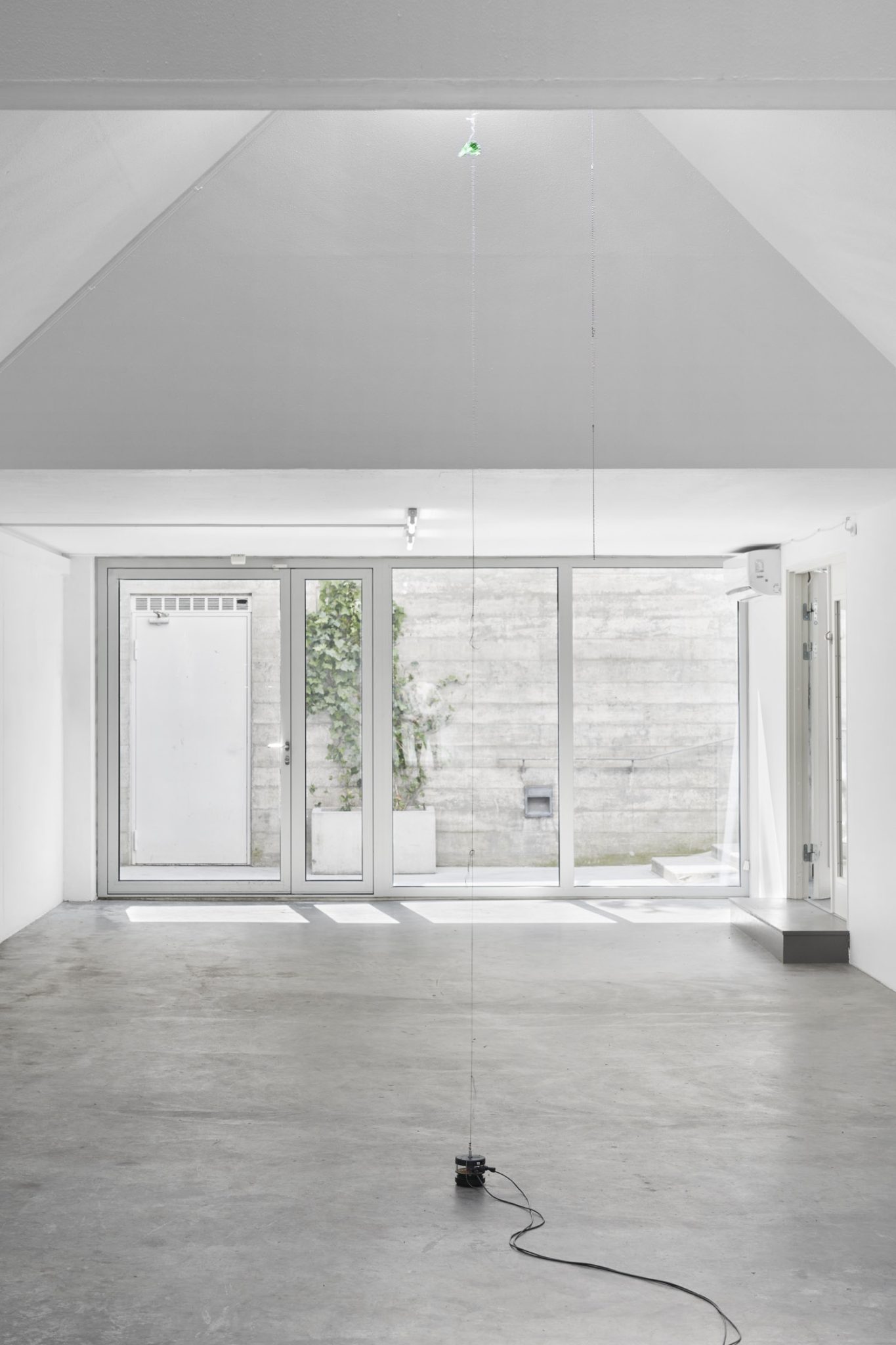 Future Mechanism Rag Plus Two Grams by Nina Canell at Simian — A minimalist gallery interior with pitched ceiling and polished concrete floor hosts an installation featuring suspended rigging equipment with green...