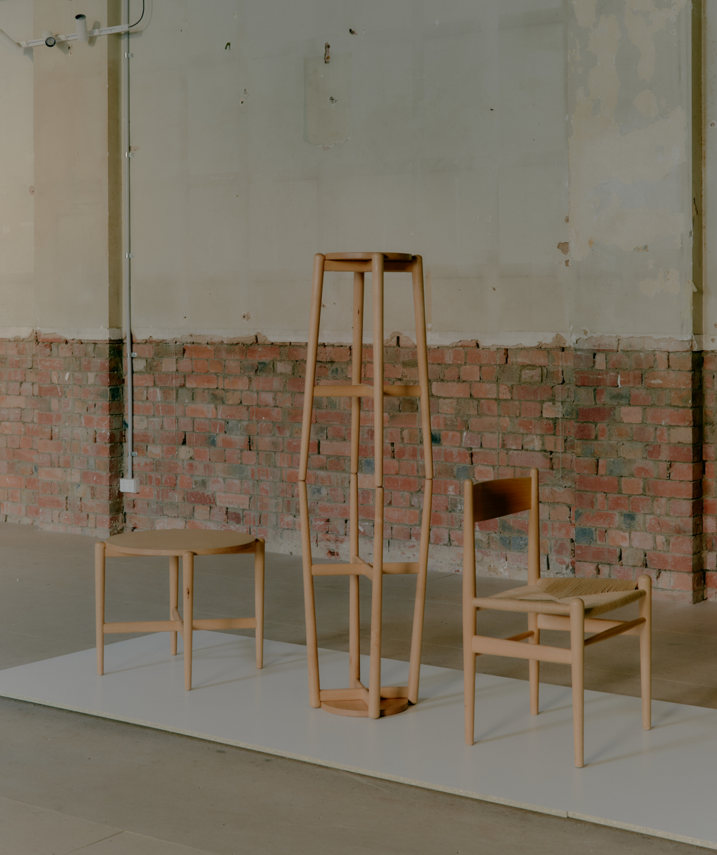 'Call Out' by DO WORKS — DO WORKS presents three timber chairs and a tall timber structure arranged on a white platform within an industrial gallery space.