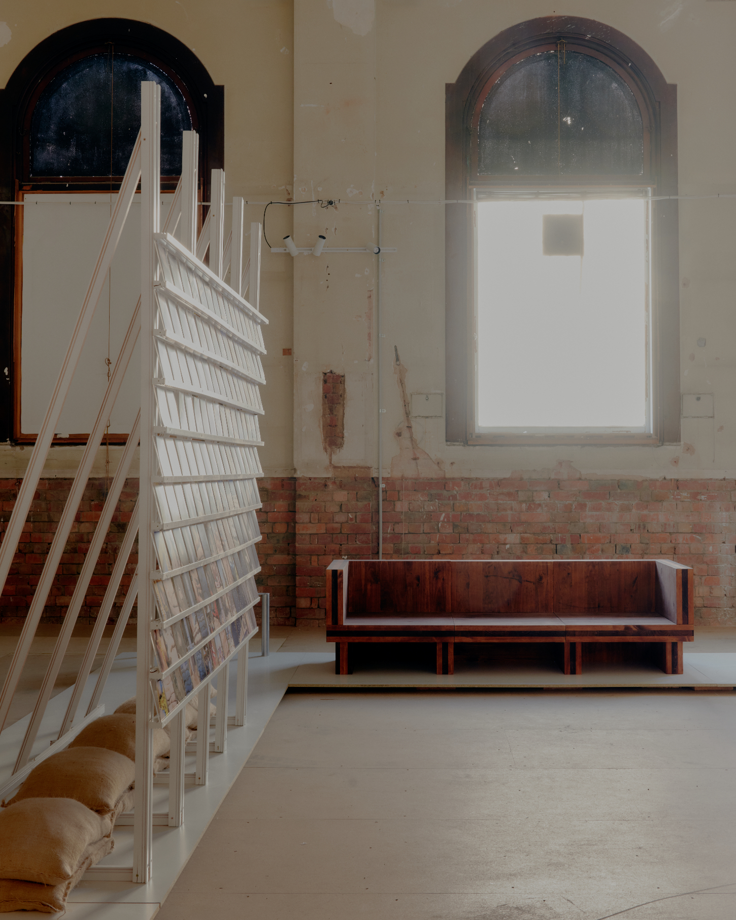 'Call Out' by DO WORKS — Within a raw gallery interior, a bespoke timber bench with burgundy upholstery sits centered against exposed brick, positioned beneath an arched window with...