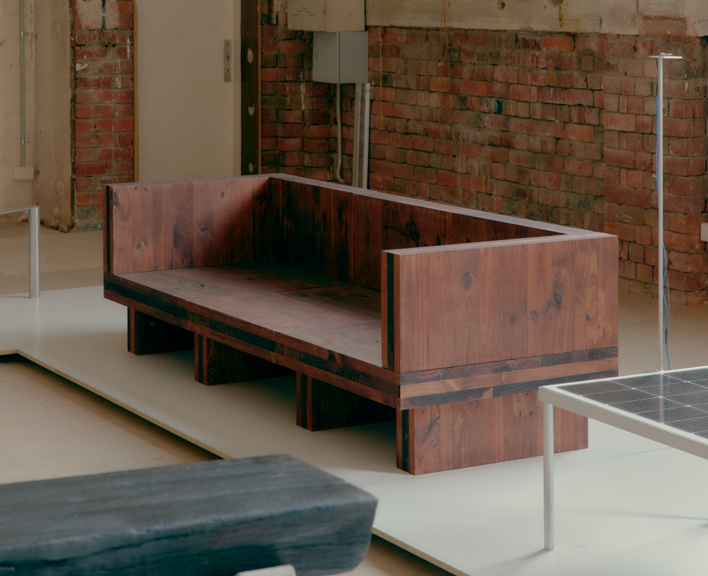 'Call Out' by DO WORKS — A timber bench with stacked wooden blocks forming an irregular base sits on a polished concrete floor against an exposed brick wall.