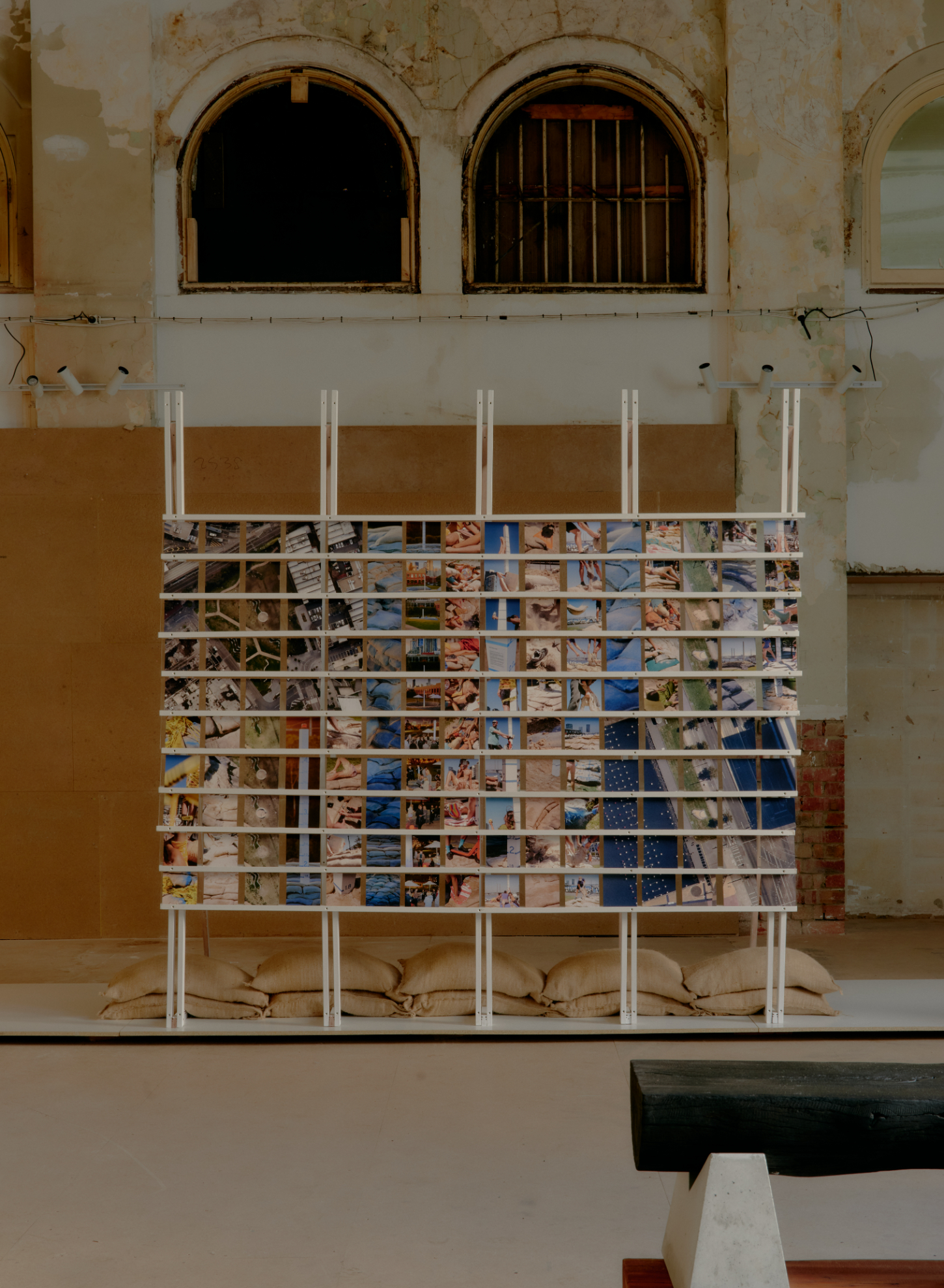 'Call Out' by DO WORKS — A modular shelving structure constructed from white steel poles supports a grid of small photographs arranged in horizontal rows, interspersed with white...