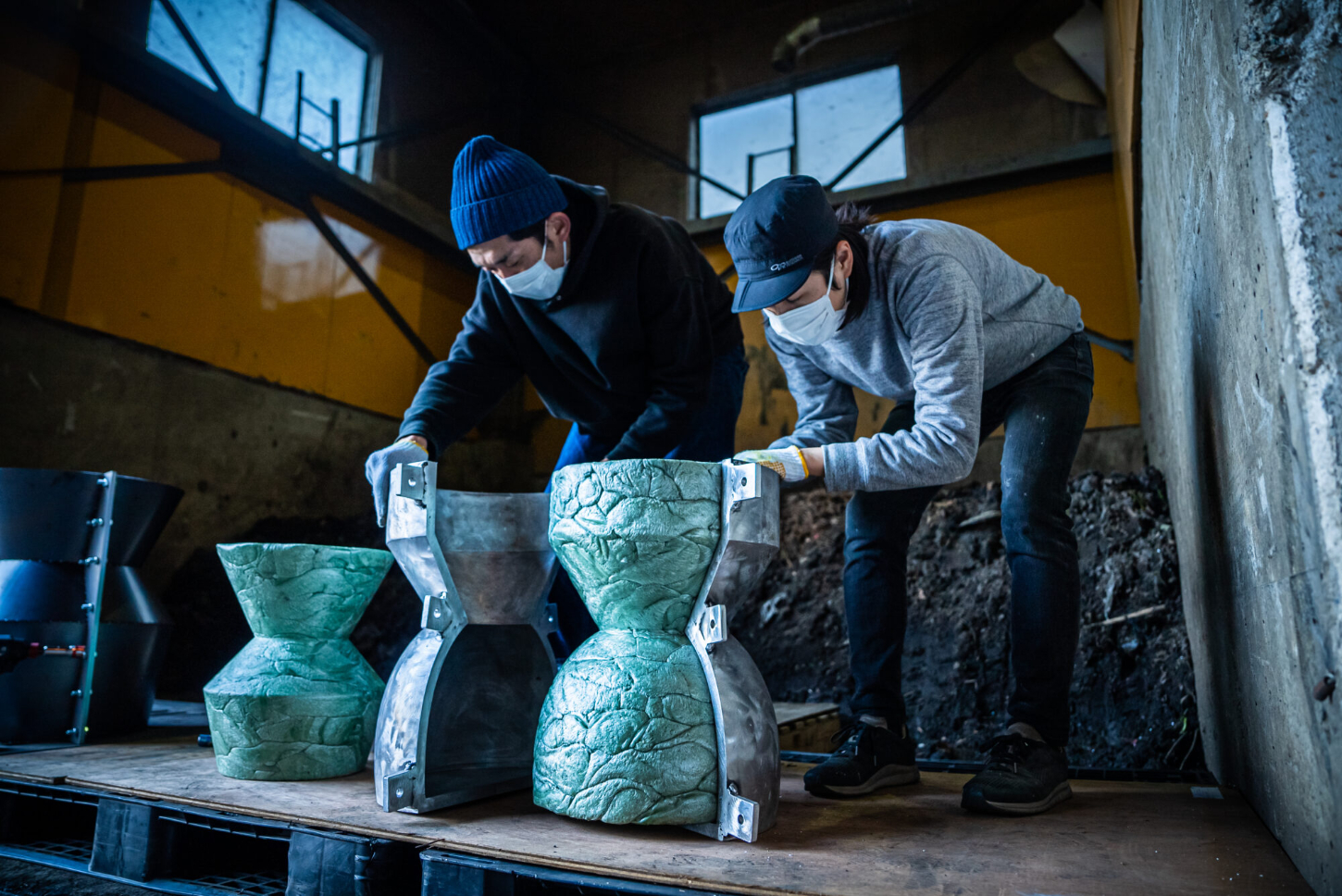 Refoam by We+ — Two workers in protective masks and beanies examine cast foam architectural capitals with carved leaf-relief ornament in an industrial workshop.