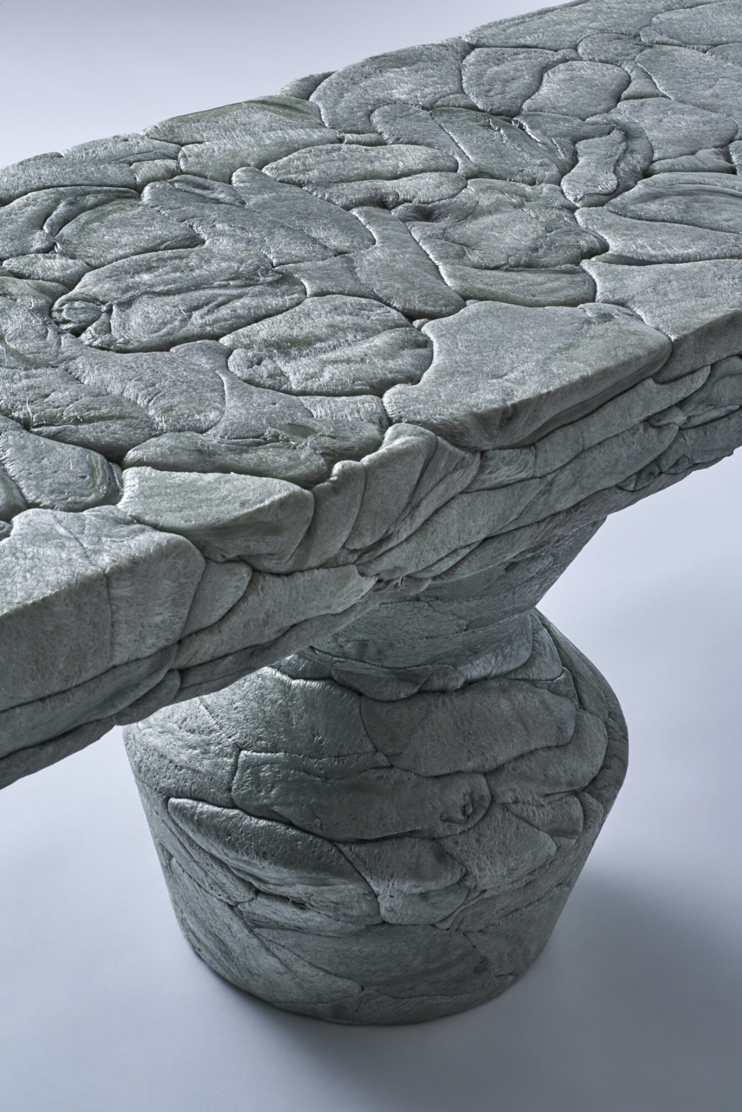 Refoam by We+ — A cylindrical architectural capital carved from concrete, its upper surface densely covered with overlapping leaf or scale motifs that create pronounced...