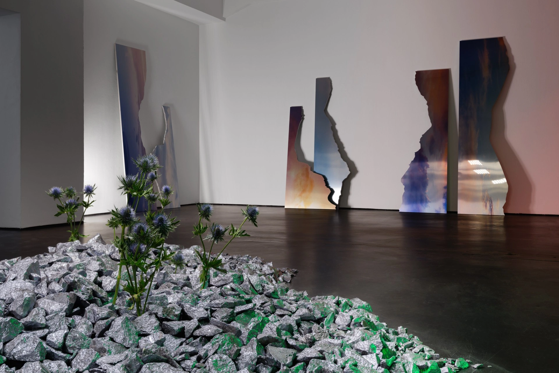 Anima Atman by Troika at Max Goelitz — The foreground installation presents a crushed mineral aggregate bed in pale grey, scattered with fragments of green-tinted stone, from which living thistle...