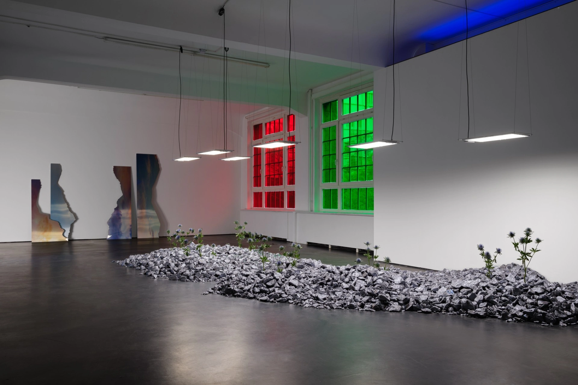 Anima Atman by Troika at Max Goelitz — A gallery interior presents a constructed landscape of crushed grey stone and mineral aggregate scattered across dark flooring, punctuated by living thistle...