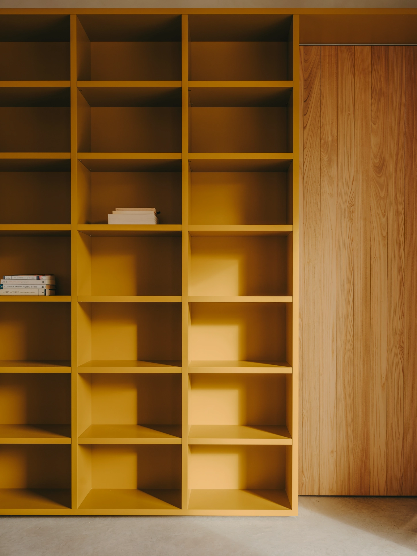 Casa TD by Skye Maunsell, Jordi Veciana and Juan Gurrea Rumeu — A gridded storage system occupies the full wall, its 3x8 compartmented structure rendered in ochre-yellow with recessed shelving creating shadow patterns...