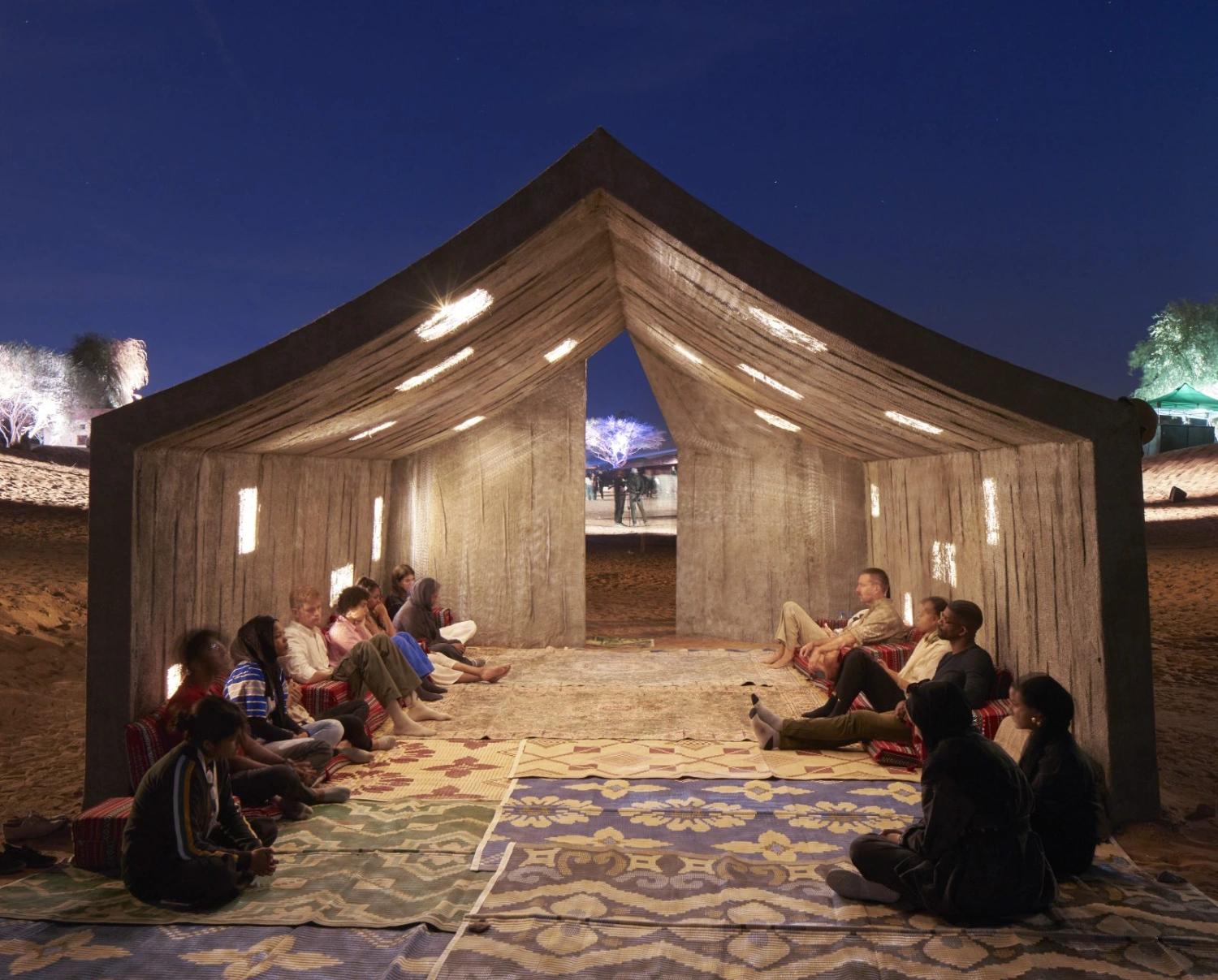 Concrete Tent by DAAR — DAAR's timber structure houses a gathering of figures seated on patterned carpets, illuminated by linear window slots that cast golden light across the...