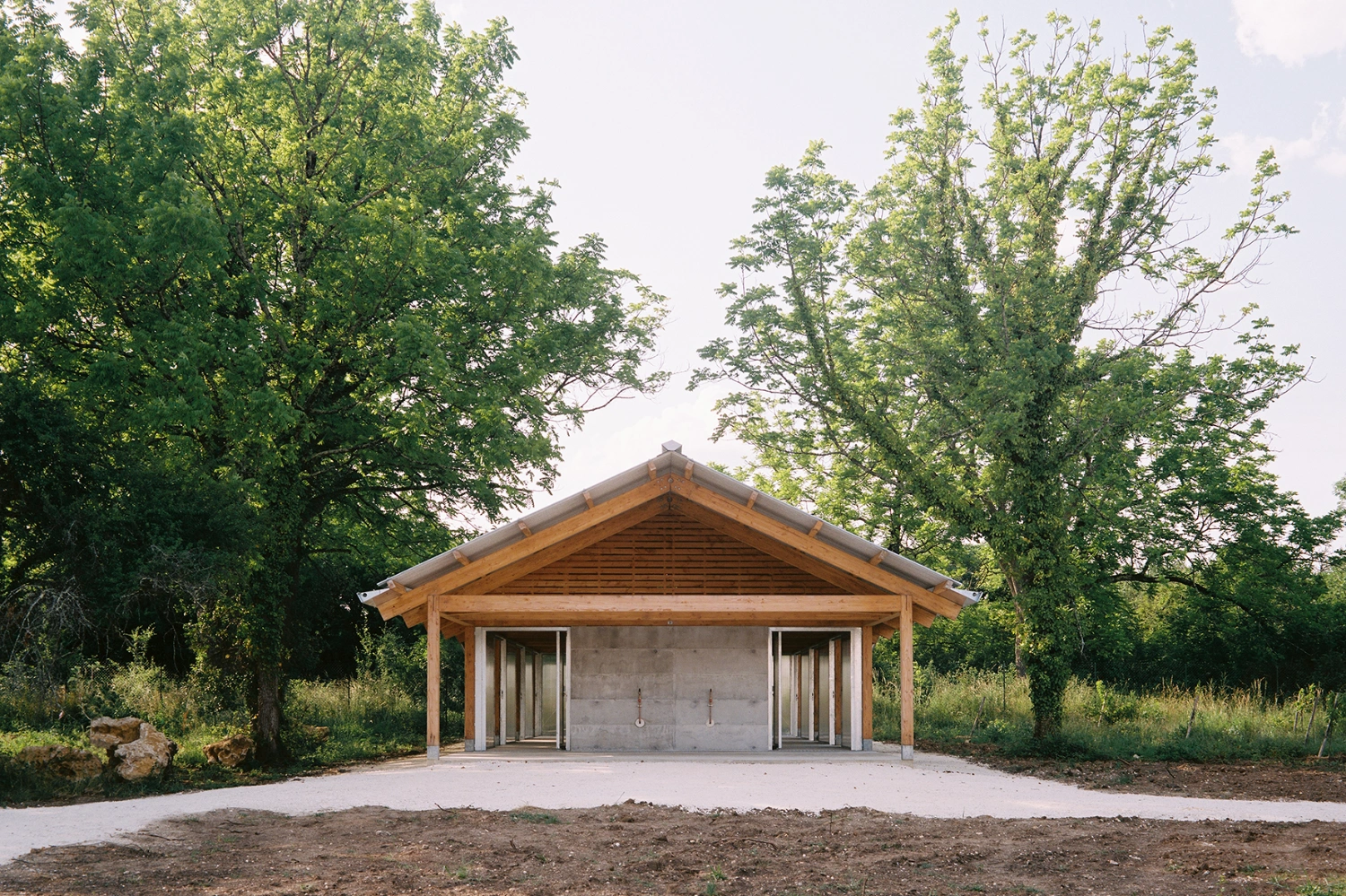 Garden Pavilion by Esnard Sanz Architectes — A timber-framed pavilion with a pitched gabled roof and exposed horizontal slat detailing in the pediment stands centered within a clearing of mature...