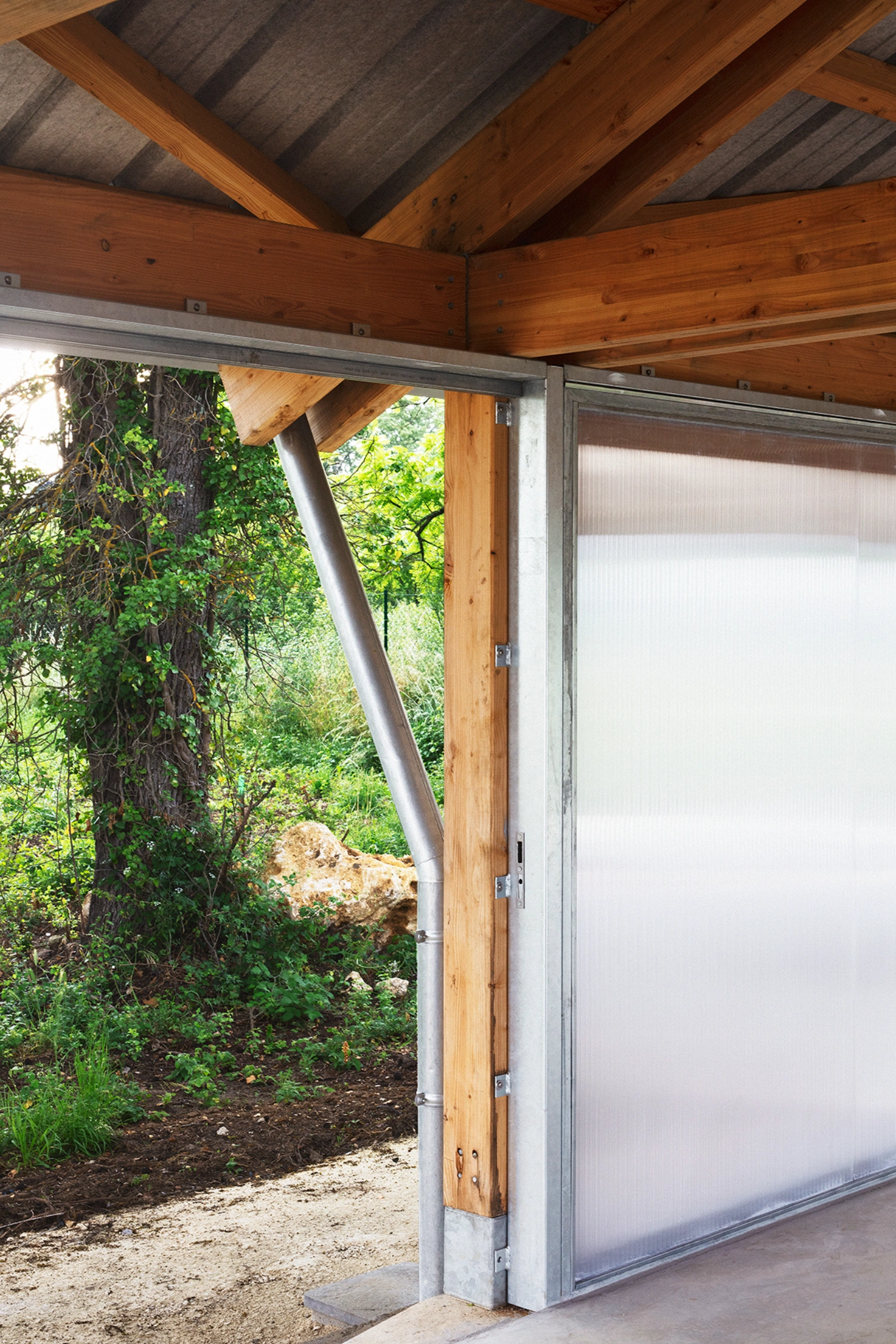 Garden Pavilion by Esnard Sanz Architectes — A timber-framed garden pavilion with a gabled roof opens onto lush vegetation through tall sliding glass doors mounted in a concrete rear wall.